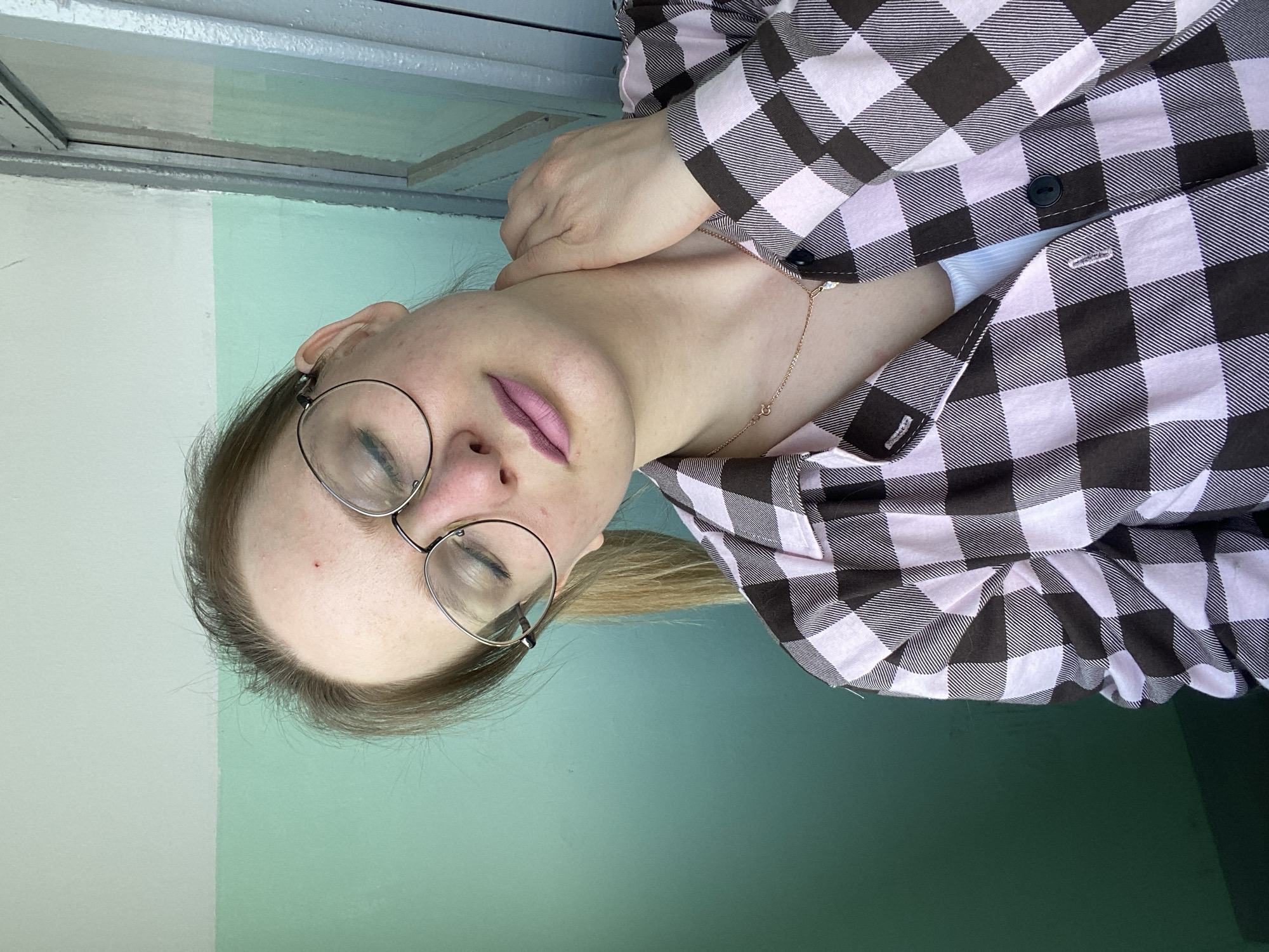 A woman with fair skin and blonde hair is positioned with her head tilted to the left of the frame. She is wearing round, wire-rimmed glasses and has her eyes closed. Her lips are colored with a pink lipstick. She is wearing a pink and black checkered flannel shirt. Her left hand is visible near her neck. A thin gold necklace adorns her neck. The background consists of two vertically divided colors: a pale white on the left and a mint green on the right. Above these colors, there is a section with a gray metal trim and a white panel. The location is Unknown, Unknown.