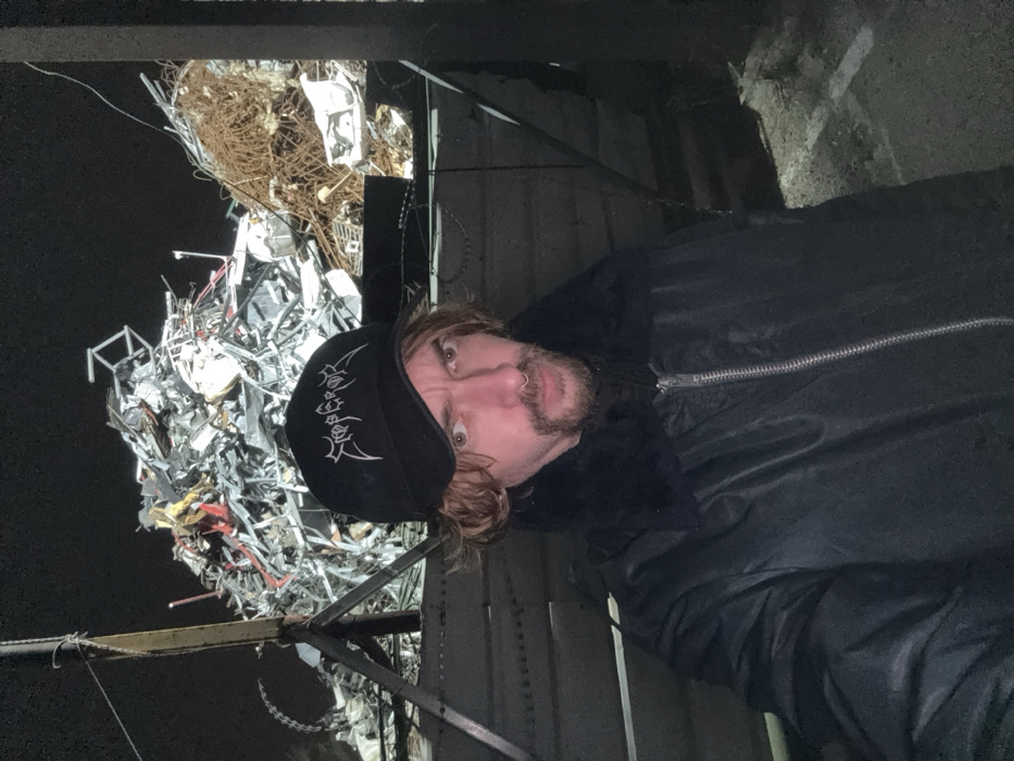A person wearing a black hat and jacket stands in front of a junkyard at night. The junkyard contains scrap metal, wires, and other debris. The person has light-colored hair and a goatee. They look directly at the camera with a curious expression. The lighting is dim, highlighting the person and the pile of junk behind them.