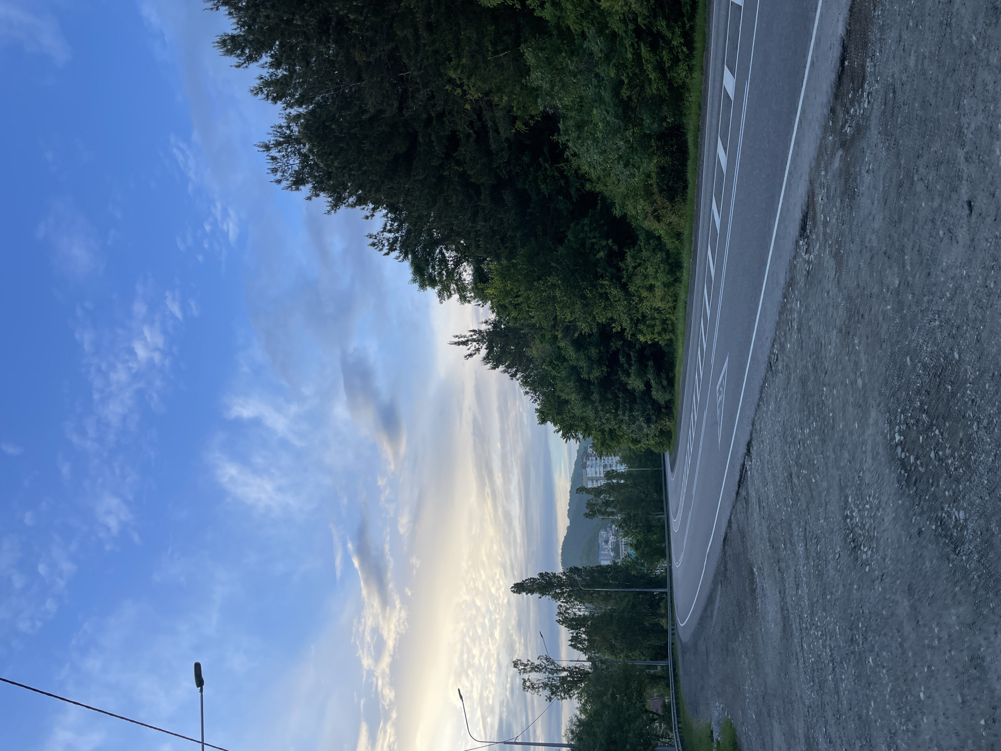 The image presents a view in Olginka, Russia, featuring a road with marked lanes curving towards the right. Dense trees line the edge of the road, transitioning into sparse trees with street lamps further down. The sky exhibits a gradient of blue, transitioning into white clouds. In the distance, there are buildings located on a hill. The foreground consists of a gravel shoulder next to the asphalt road.