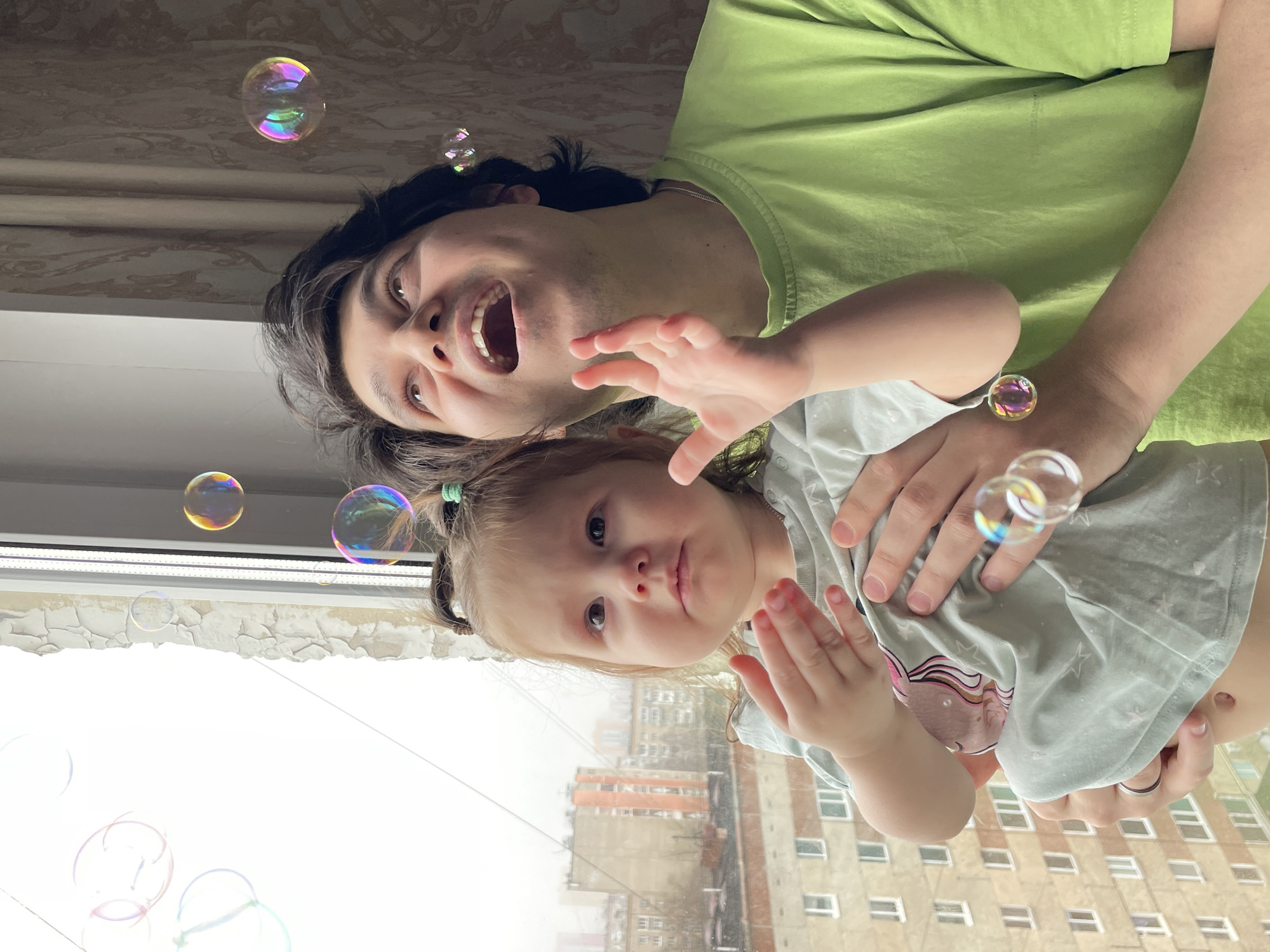 An overhead-angle photograph captures a man and a young child playing with soap bubbles indoors, against the backdrop of an urban landscape in Izhevsk, Russia. The image is oriented vertically, with the subjects appearing from below.
The adult, a man with dark hair, occupies the upper part of the frame, wearing a light green t-shirt. He has a wide-open mouth, suggesting laughter, surprise, or excitement, and his eyes are wide and focused. A silver ring is visible on his left ring finger.
Held by the man is a young child, likely a girl, with light brown hair styled in pigtails secured with green ties. She wears a light grey or green t-shirt with a partially visible graphic design. Her hands are raised and slightly open, with a reddish mark on one hand, and she has a neutral, perhaps slightly curious or pouty expression.
Numerous iridescent soap bubbles float throughout the scene, reflecting vibrant colors like pink, purple, yellow, and blue.
The setting combines an indoor space with an outdoor view. Above the subjects, a patterned, light-colored ceiling or wallpaper is visible. Through a large window, an exterior view of a residential area is seen, characterized by multi-story apartment buildings made of various shades of brick and concrete. The sky beyond the buildings appears bright and overcast, indicating daytime. The window frame is white, and the wall just beneath it shows signs of peeling paint or plaster.
The activity is a playful moment between the man and child, centered around the soap bubbles, conveying a sense of joy and engagement. No visible text is present in the image.