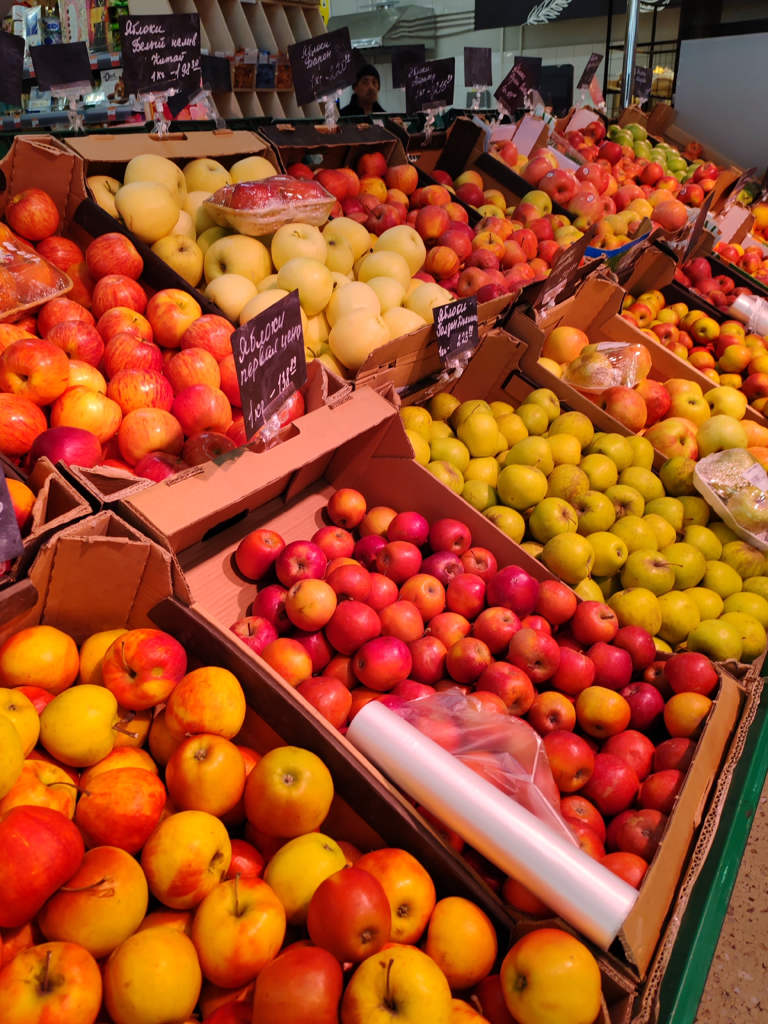 The image shows a display of apples in a grocery store, likely in Ust-Ilimsk, Russia. A variety of apples in cardboard boxes are stacked on shelves, with different colors and types displayed, including red, yellow, and golden apples. Some of the apples are wrapped in plastic. Signage in Russian indicates the apple types and prices per kilogram. Rolls of clear plastic wrap are also visible. An individual is blurred in the background, possibly a store employee.