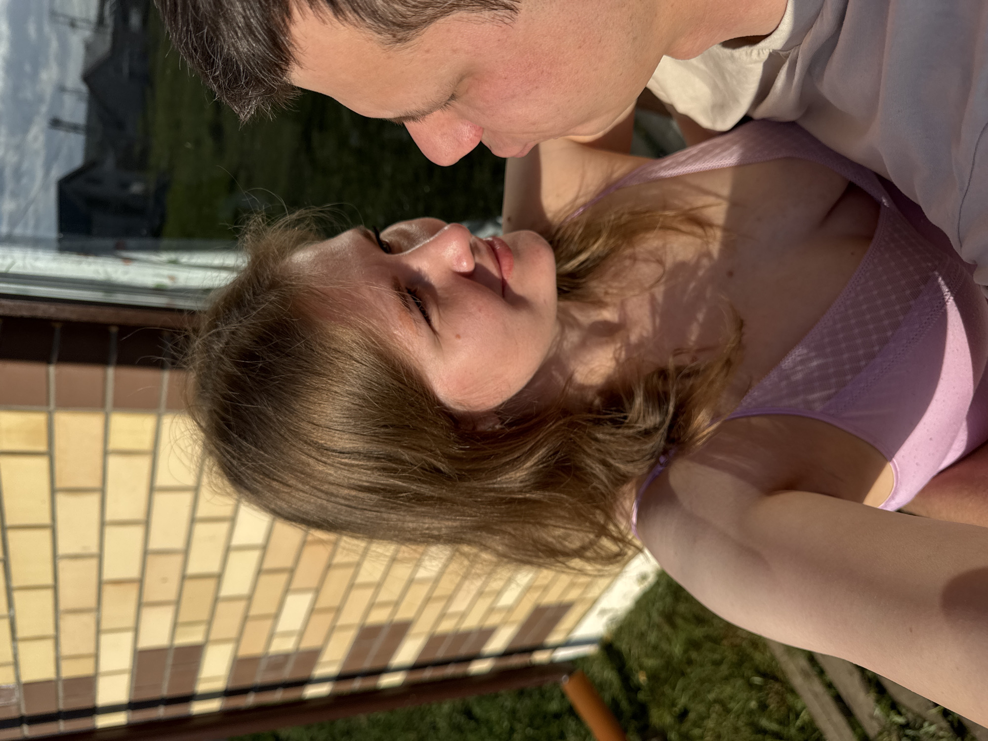A light-skinned couple appears to be kissing outside a building. The man has dark hair, and is wearing a light-colored t-shirt. The woman has light brown hair and is wearing a light purple tank top. They are standing near a window with the reflection of a building. A brick structure is visible nearby. Outside of the structure, grass and foliage are seen. This image was taken in the city of Ферма, Russia.