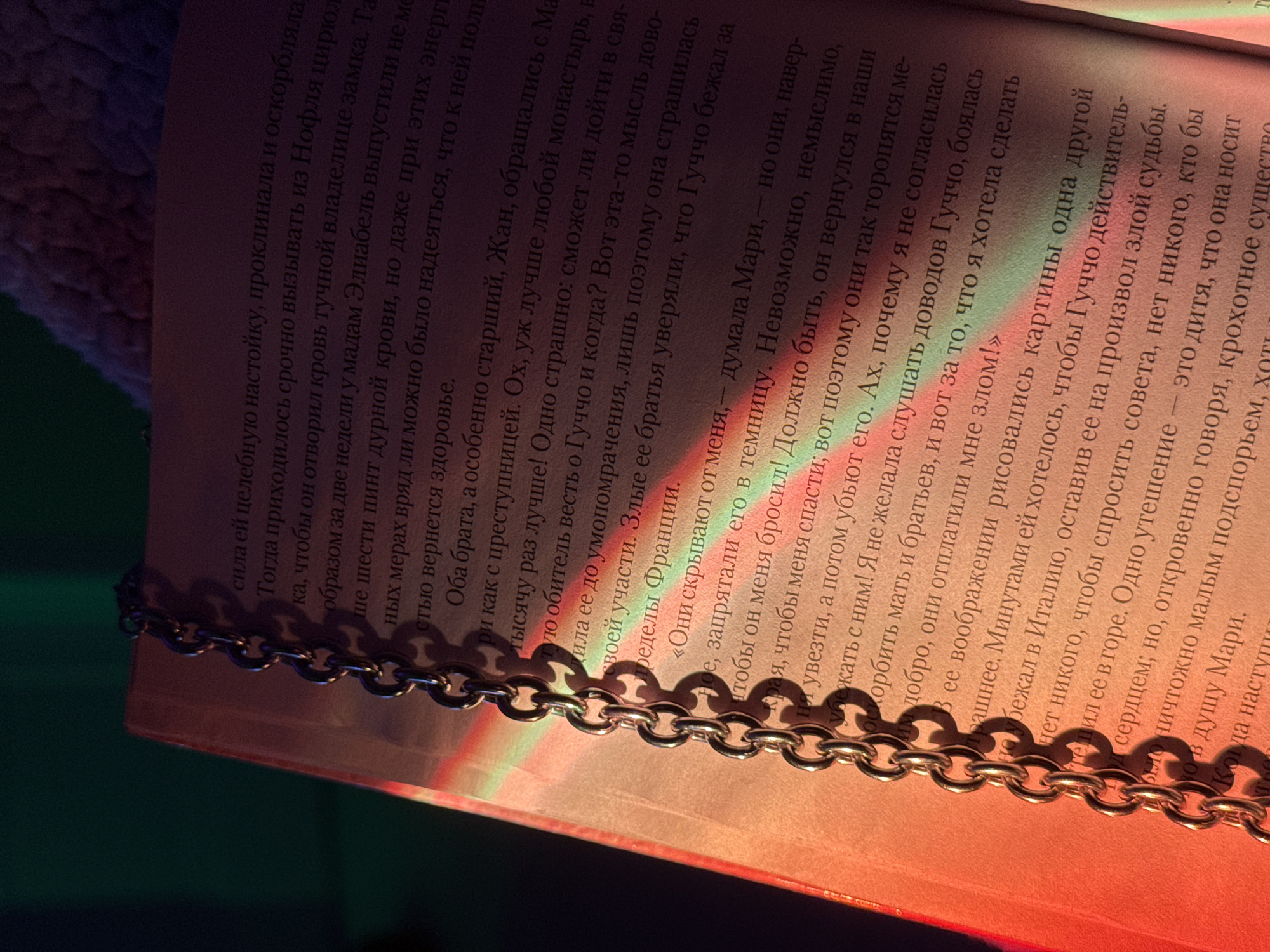 Here is a detailed description of the image:
**Scene:** A close-up shot of a page from a book. The text is in Russian. The book has a gold chain draped on top of it. The lighting is warm, and the text appears to be from a novel or story, focusing on characters named "Мари" (Mari) and "Гуччо" (Guchcho).
**Text/Content:** The visible text passages describe the character Mari, her thoughts, and her situation. She reflects on Guchcho's possible fate, her brothers' actions, and her own regrets. The narrative indicates a sense of betrayal, fear, and confinement. The passage also mentions a "дитя" (child) that she carries.
**Objects:**
* Book
* Gold Chain
* Russian text
**Environment:** The environment is implied through the text. It describes possible locations like a "темницу" (dungeon/prison) or France. The text also evokes emotions of fear and confinement.