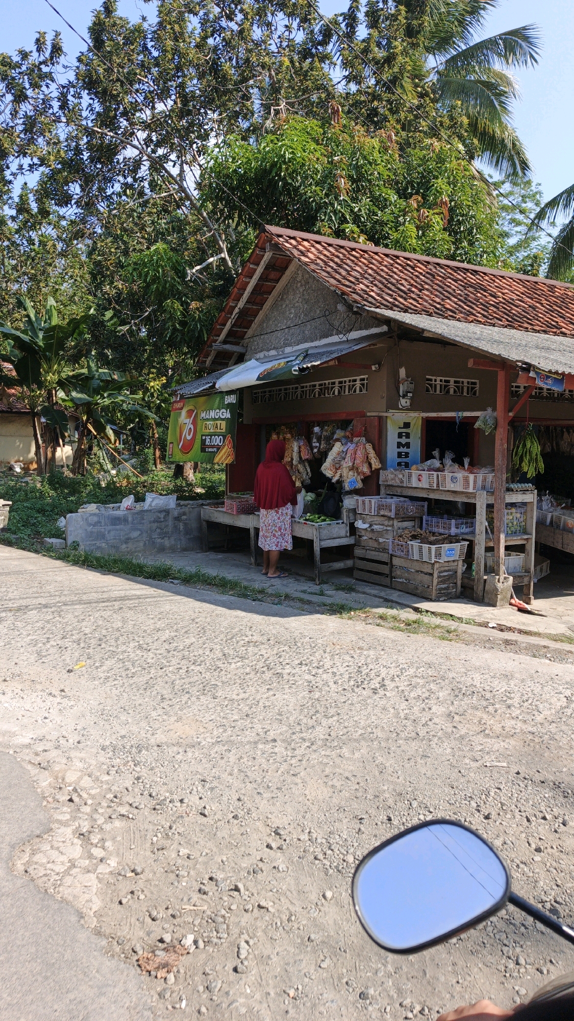 A full shot captures a small roadside stall in Desa Batukaras, Indonesia, under a clear blue sky. The stall, constructed with concrete and wood, has a reddish-tiled roof and is stocked with various goods. To the left, a woman wearing a red headscarf and a floral-patterned dress stands facing the stall, appearing to interact with its offerings. She is positioned in front of a display of what appear to be green fruits. In front of the stall, a wooden table holds more produce, including several green items. Behind the woman, a prominent yellow sign with red lettering advertises "MANGO ROYAL" at a price of "16.000". Shelves behind the counter are laden with packaged goods and produce, including baskets filled with items and bunches of green vegetables hanging at the far right. The stall is situated next to a rough, unpaved road. Lush green trees and foliage surround the scene, with banana trees visible to the left. In the foreground, the reflection of the blue sky and power lines is visible in the side mirror of a motorcycle. The overall impression is that of a typical rural market scene on a sunny day.
