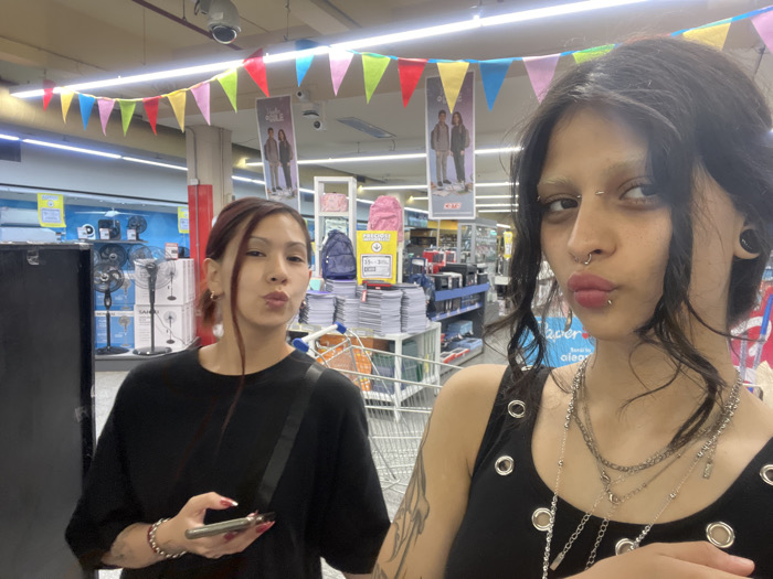 Two women pose in a store with colorful flag banners overhead. The woman on the right wears a black tank top with metal rings, several necklaces, and facial piercings. She has dark hair and light-colored eyebrows. The woman on the left wears a black t-shirt and is holding a phone. Both women are making a kissing face. A shopping cart and shelves stocked with items are visible in the background. Fans in boxes and promotional posters are also seen in the store.