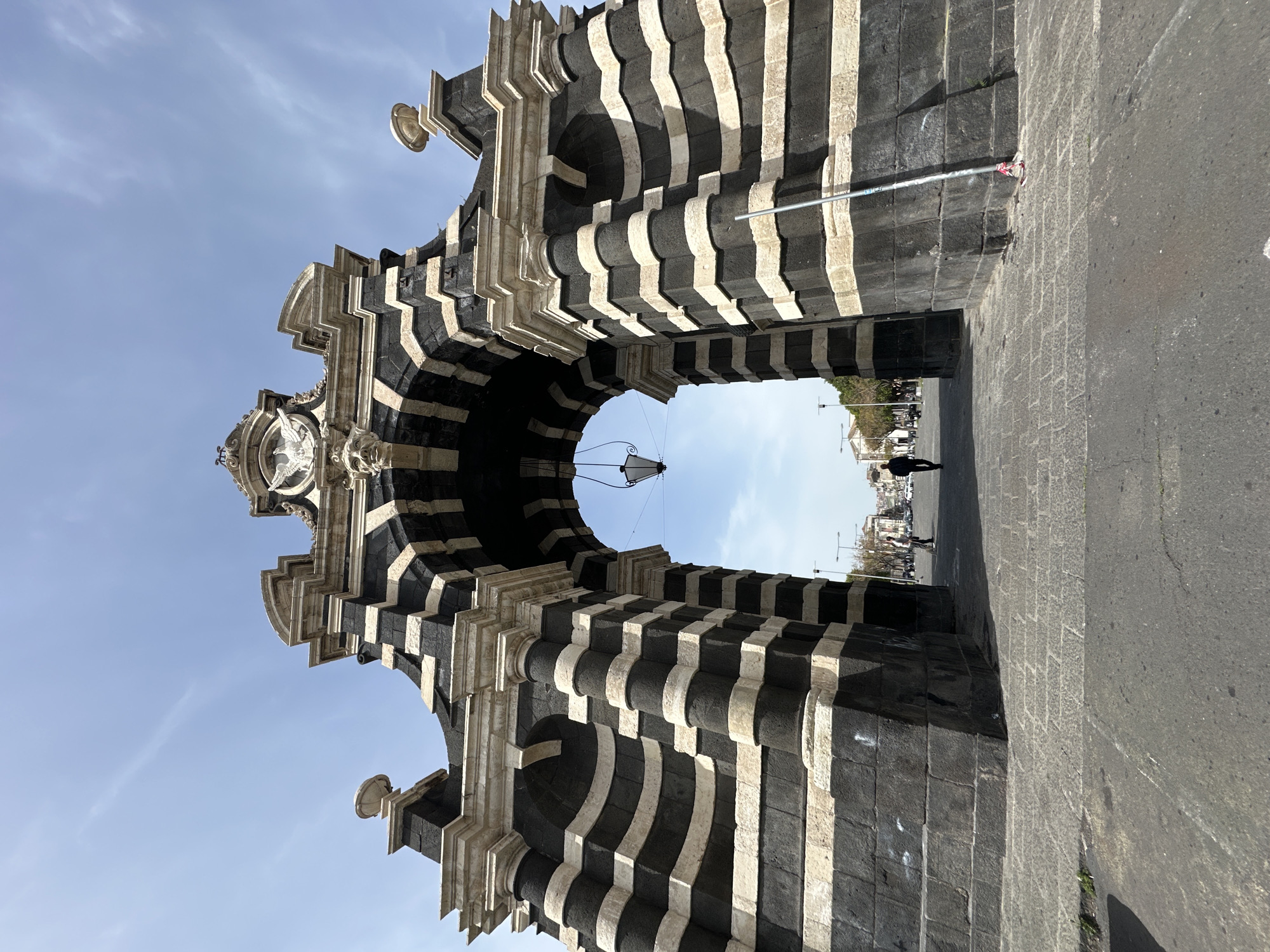 The image shows the Porta Garibaldi, a triumphal arch located in Catania, Italy. The arch is constructed with alternating layers of black lava stone and white limestone, giving it a striped appearance. The archway frames a view of a street with pedestrians and buildings in the distance. A lamp hangs from the center of the arch. The sky is blue with a few clouds. The street is made of stone.