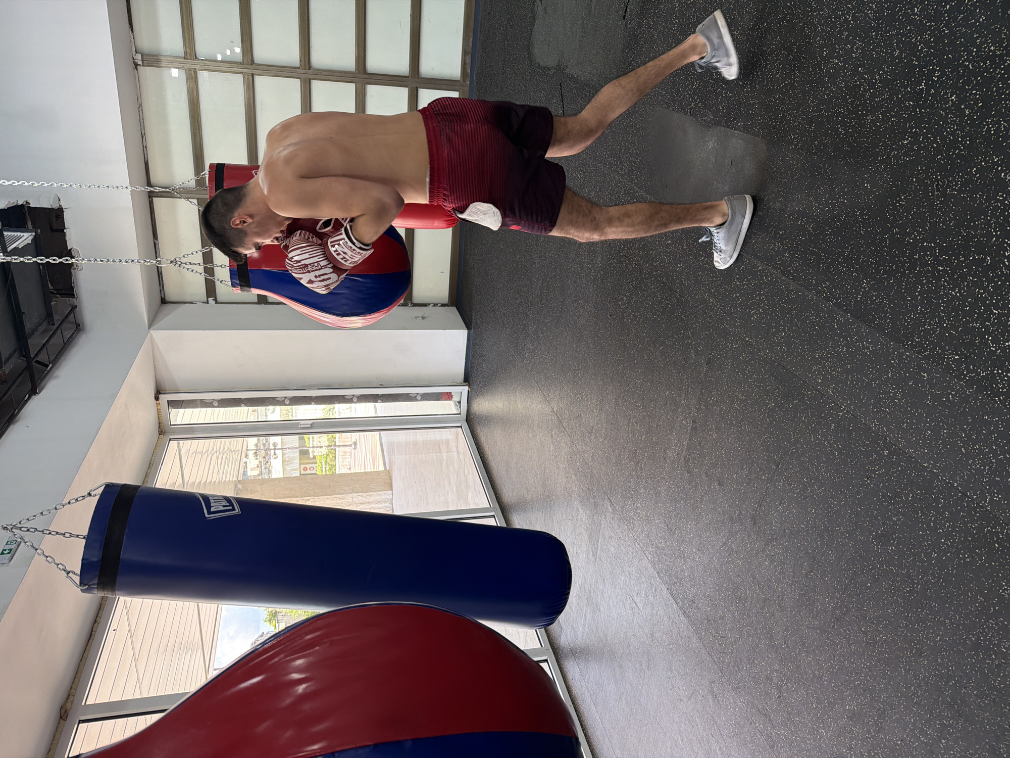 A shirtless man wearing red and black striped shorts and grey sneakers is training with a punching bag in a gym. He is wearing boxing gloves and is in a fighting stance. The gym has large windows and a black rubber floor. Several punching bags of various sizes and colors are visible. The scene is set in a modern gym with good lighting.