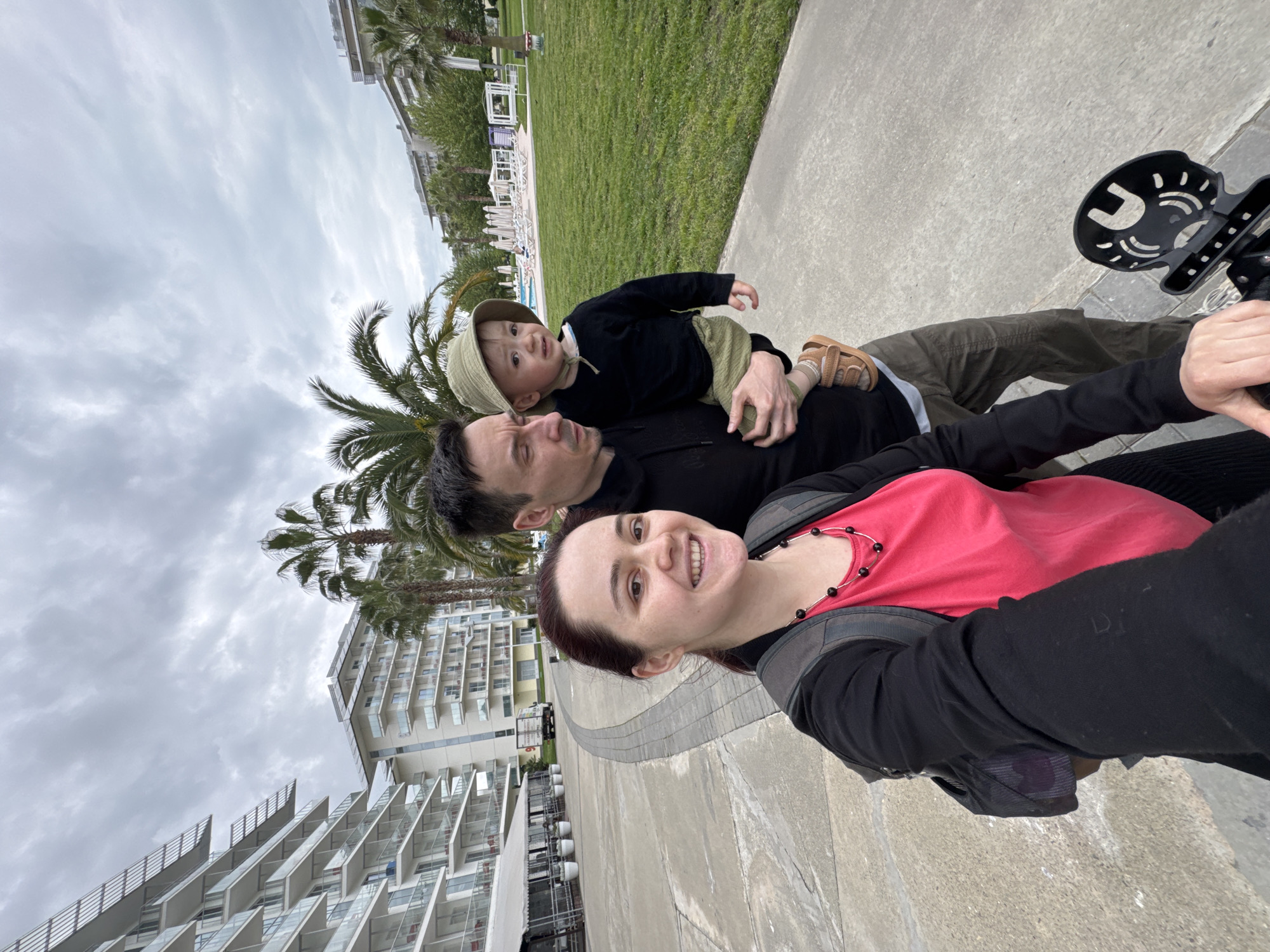 The image shows a family of three posing for a selfie in Sochi, Russia. The woman taking the photo is wearing a pink top and a black jacket, with a backpack over her shoulders. Her hair is pulled back. The man is lying on the ground, wearing a black shirt and green pants. A young child is lying on top of the man, wearing a black shirt and a green hat. The background includes palm trees, modern buildings, a grassy area, and a cloudy sky. There is a child's bike seat visible.