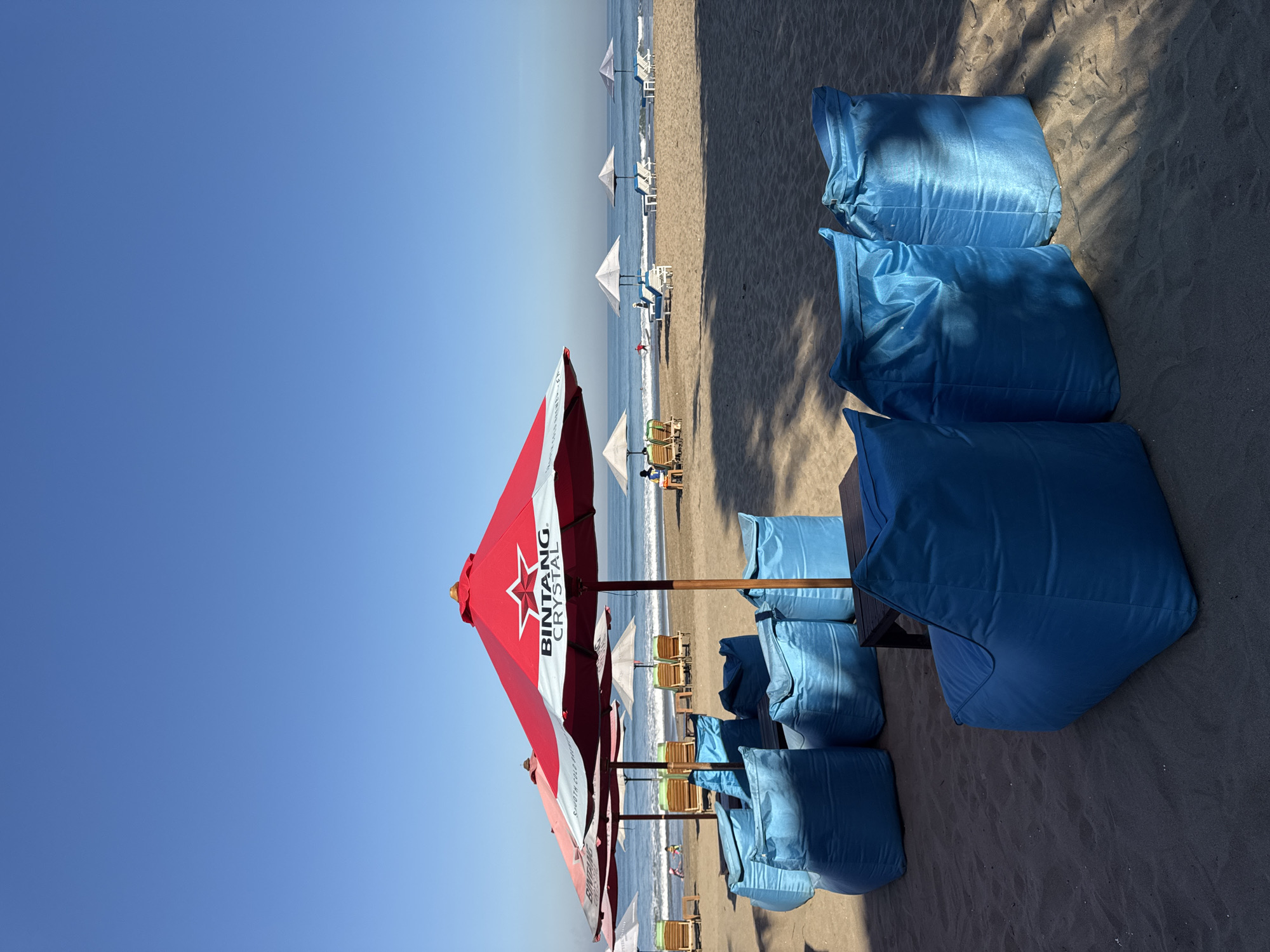 The image depicts a beach scene, likely in Kuta, Indonesia. On the sand, there are rows of blue beanbag chairs arranged near red and white umbrellas branded with "Bintang Crystal," likely indicating an advertisement for a local beer. The beach also contains additional sunshades and lounge chairs. In the background, there is an ocean and clear blue sky. There are people visible on the beach, suggesting a leisure or vacation setting. The image is oriented vertically.