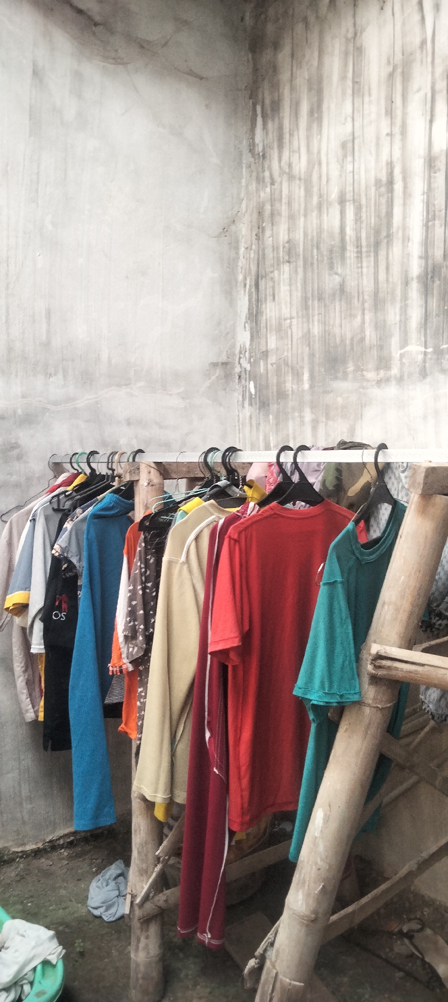 This image captures a clothesline filled with various garments hanging from hangers against a textured concrete wall in what appears to be an outdoor setting. The clothesline itself is supported by a rustic wooden structure made of bamboo poles. The clothing items include t-shirts and long-sleeved shirts in a range of colors such as red, blue, beige, and teal, some with intricate patterns. On the ground below, a pile of light blue fabric is visible. The overall scene suggests a domestic or utilitarian space, possibly a backyard or alleyway in Antirogo, Indonesia, given the context.