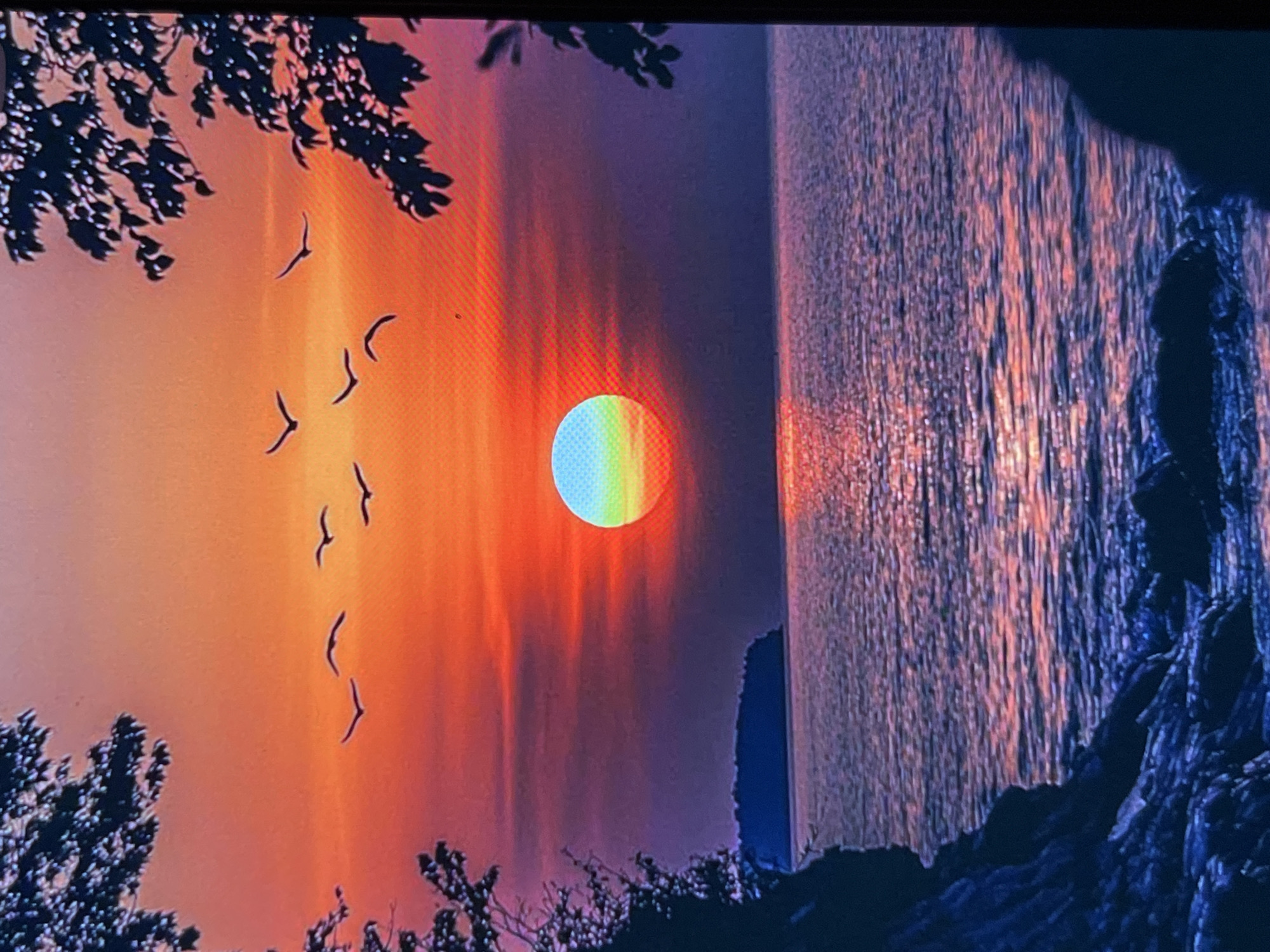 This media file depicts a scenic sunset over a body of water, with a rocky coastline in the foreground. The sun, positioned in the center of the frame, appears as a vibrant orb with a rainbow-like spectrum. Silhouettes of trees frame the top and bottom edges of the image, and a flock of birds is captured in mid-flight against the colorful sky. The water reflects the intense colors of the sunset, creating a shimmering effect. The overall mood is tranquil and awe-inspiring, evoking a sense of natural beauty. The image is a photograph or digital artwork, likely representing a landscape scene. No visible text is present. The location context provided is Montfermeil, France, suggesting this scene may be inspired by or located in that area, known for its natural parks and scenic views.
