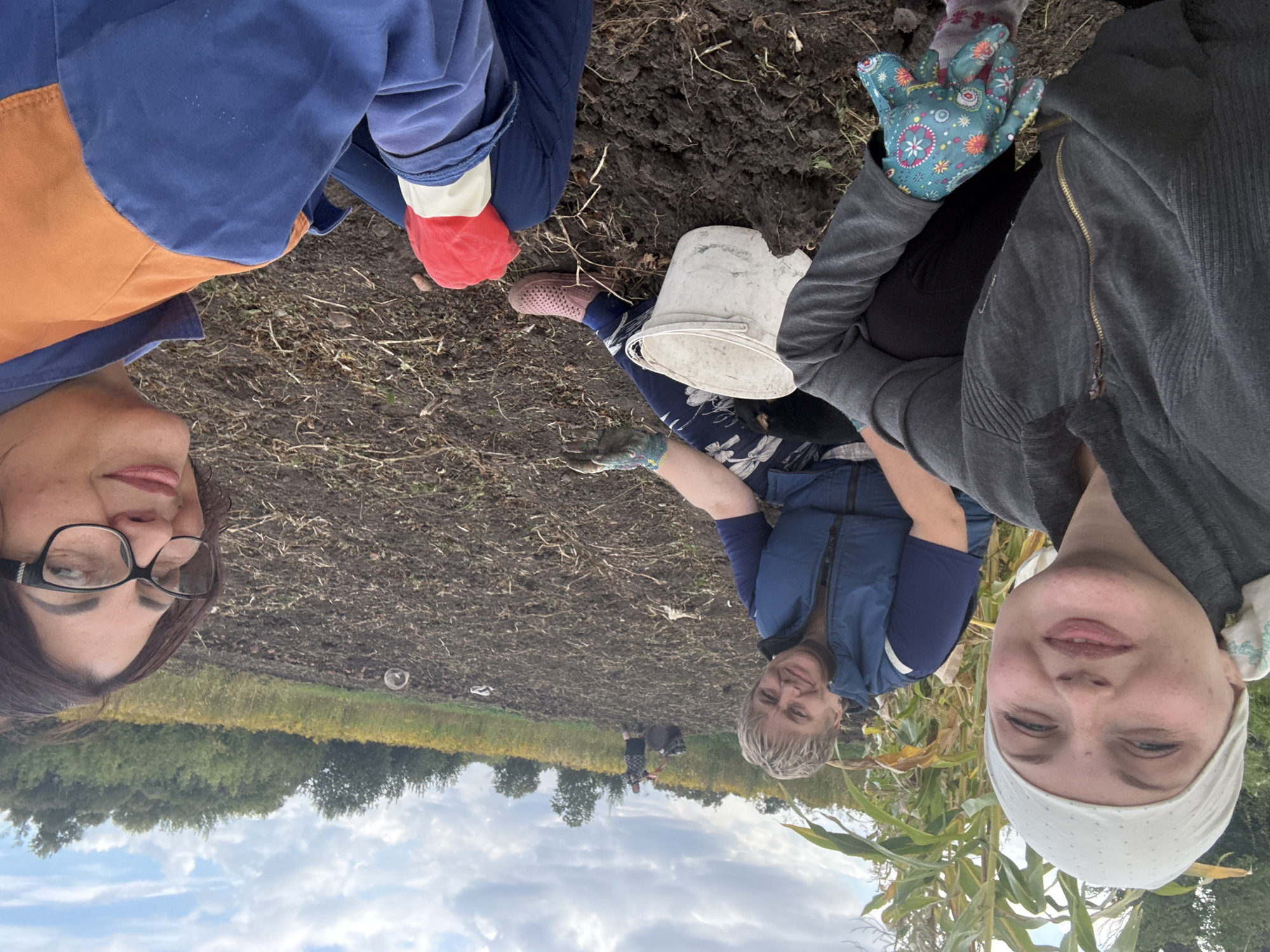 Three women are posing for a selfie in a field in Fominka, Russia. The woman in the foreground on the left is wearing a grey zip-up jacket and a white headscarf. She has a floral-patterned glove on her left hand. The woman in the middle is wearing a blue vest over a dark blue shirt and has short blonde hair. She is wearing gardening gloves and has a white bucket next to her. The woman on the right is wearing glasses and an orange and blue jacket. She has a red glove on her left hand. The field appears to be recently tilled, with brown soil and scattered plant debris. In the background, there is a person operating a tiller in a yellow field of flowers, and trees line the horizon under a cloudy sky.