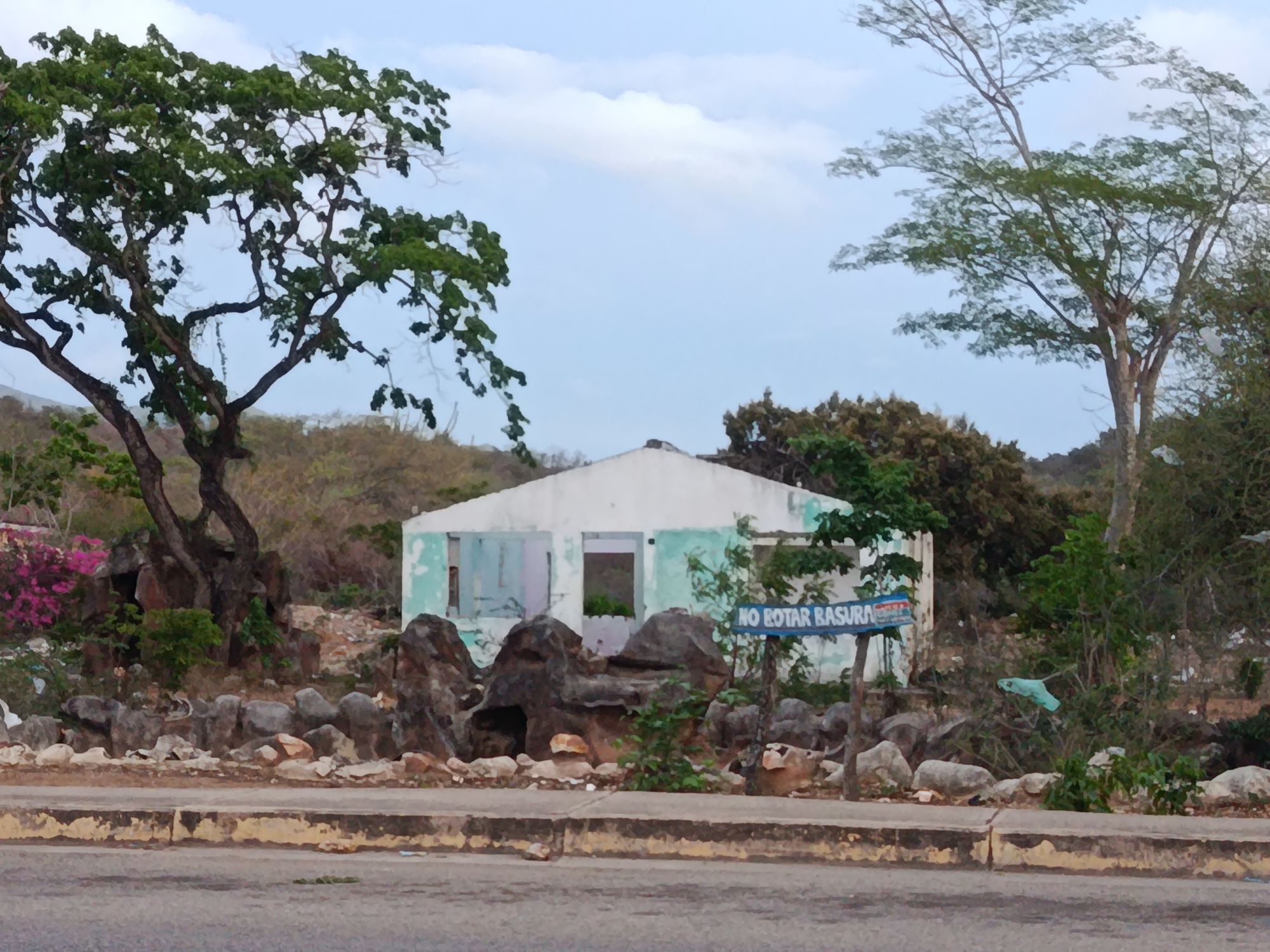 A low-angle, medium shot captures an abandoned, small, white structure with teal accents, situated in a natural, rocky landscape. The building, appearing to be a house or small business, has open doorways and windows, some covered with what appear to be shutters. In front of the building, large rocks are scattered, forming a rough border. A sign on a wooden post reads "NO ROTAR BASURA," indicating "Do Not Throw Trash." To the left of the building, a large tree with a thick trunk and branches spreads its green foliage against a cloudy sky. To the right, another tree with a more delicate canopy is visible. The ground is uneven, with sparse vegetation and some debris. The scene is bordered by a paved road in the foreground. The environment suggests a semi-rural or undeveloped area within the city of Altagracia, Venezuela.