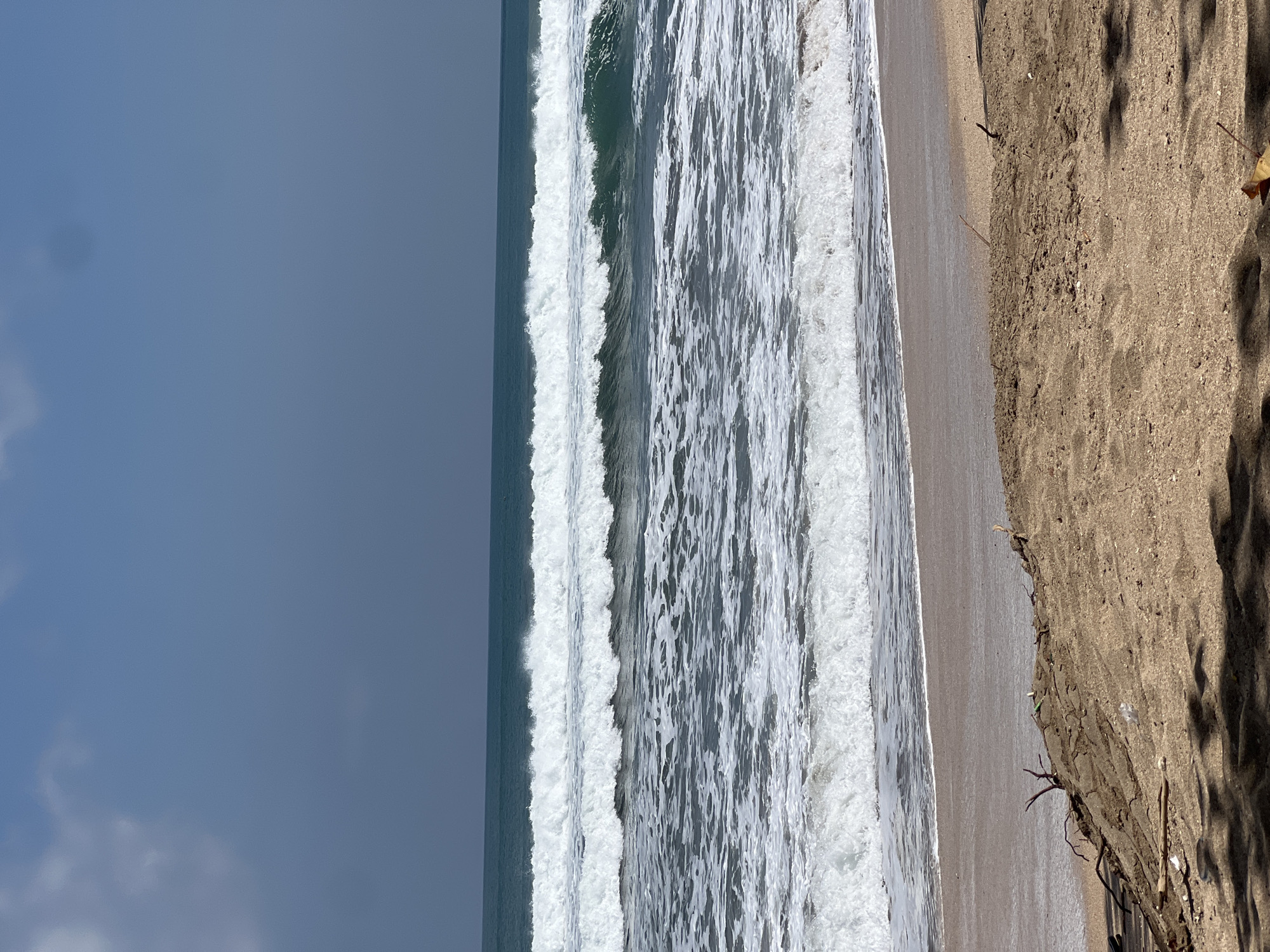 The image depicts a serene beach scene in Kuta, Indonesia, under a clear blue sky with scattered clouds. The ocean is calm, with gentle waves rolling towards the shore. The sand on the beach is a light brown color, with some debris scattered on it, including twigs and dry leaves. The foreground shows the sandy shore with some shadows from unseen trees. The middle ground is dominated by the ocean, with several rows of waves breaking. The background features a darker expanse of the ocean meeting the sky. There are no people visible in the image, and the scene appears to be undisturbed. The overall atmosphere is peaceful and natural, characteristic of a tropical beach.