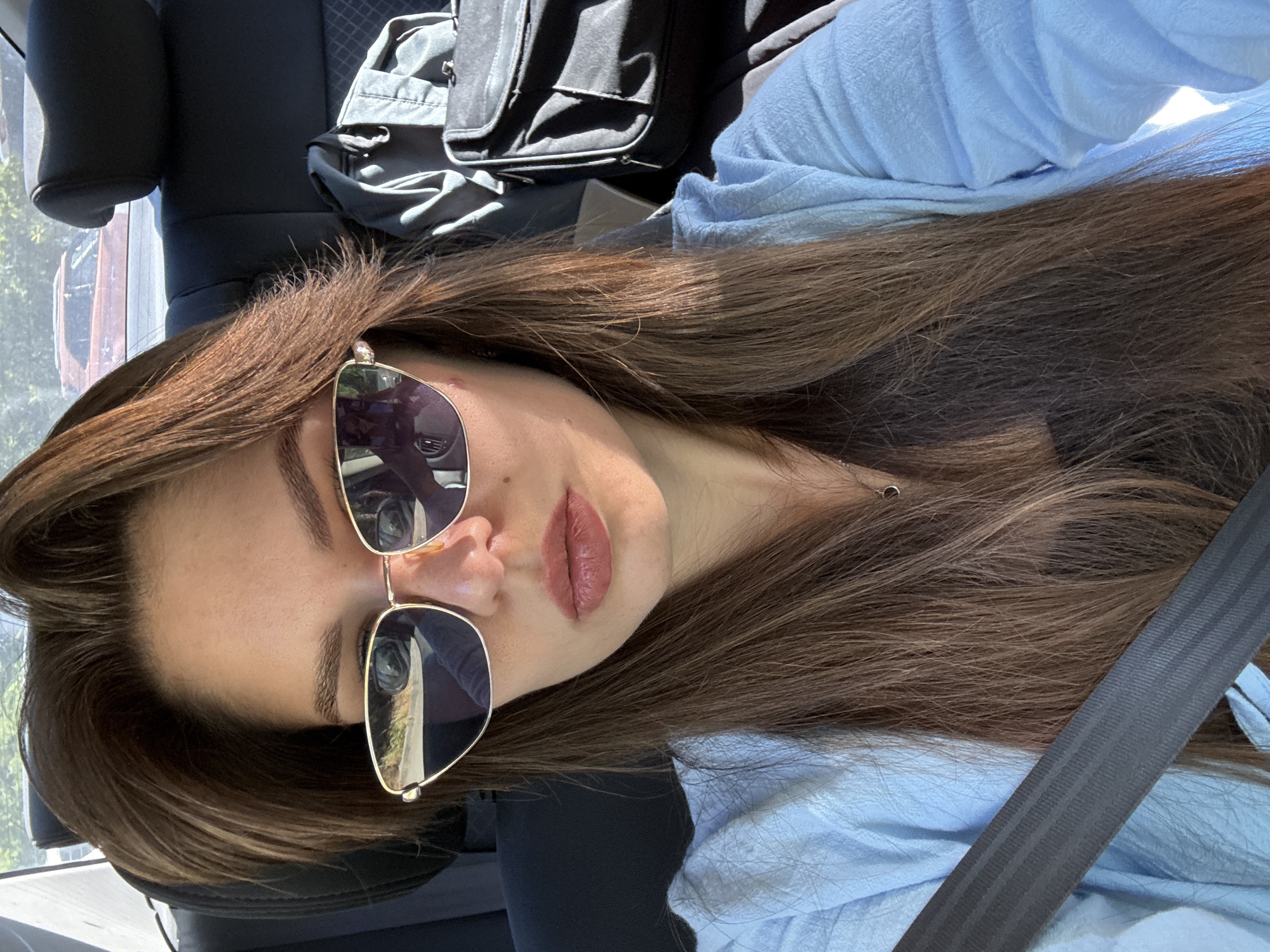 A woman with long brown hair is sitting in the back seat of a car, wearing a light blue shirt and sunglasses. She is looking at the camera, and her right hand is visible. A seatbelt is across her chest. In the background, there is a black bag and the interior of the car. Outside the car window, another car is visible. The scene appears to be in a sunny outdoor environment.