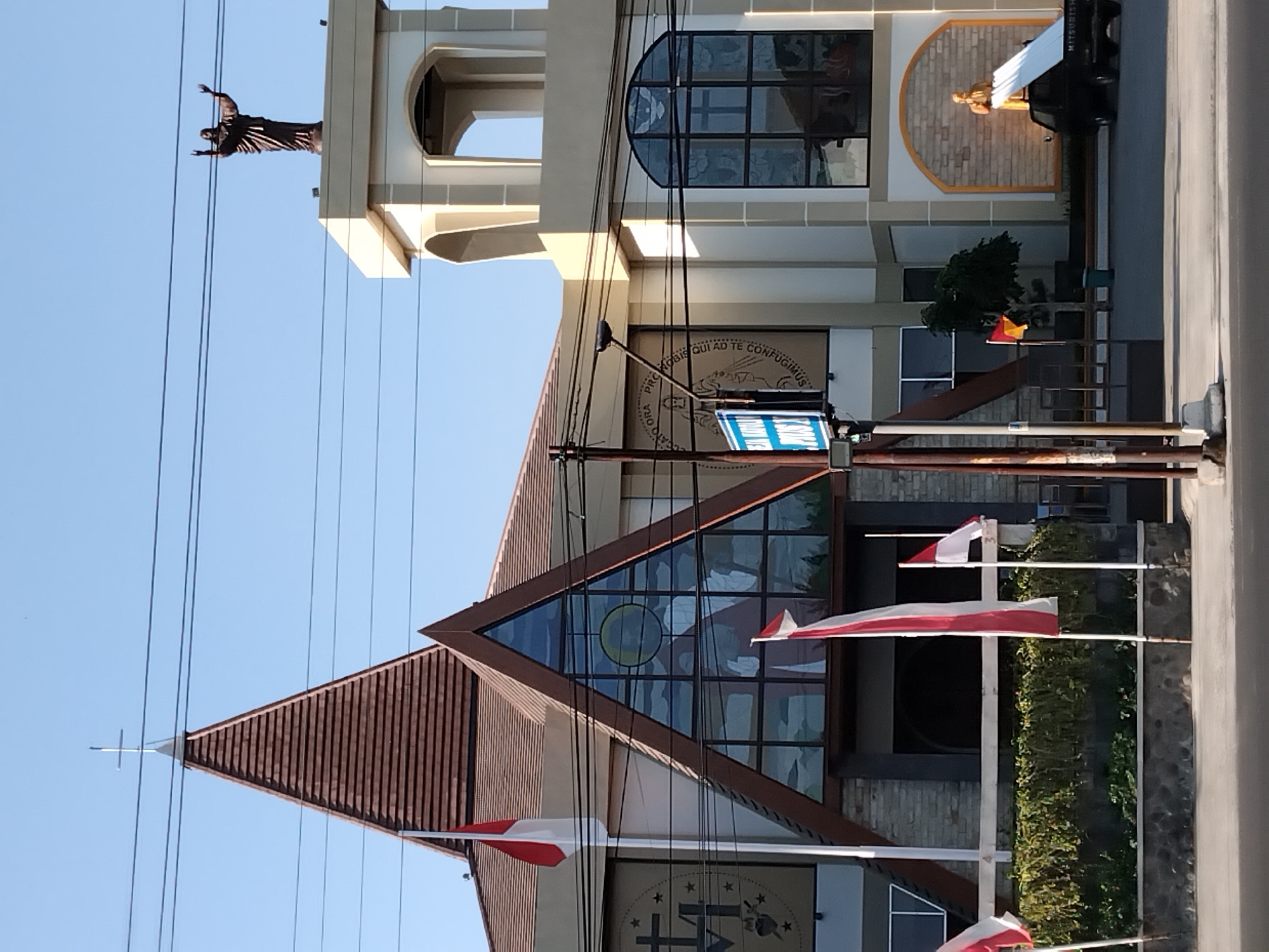 The image captures a frontal view of a church building, likely located in Karanganyar, Indonesia, due to the prominent display of the Indonesian flag. The church features a distinctive triangular facade with a large stained-glass window depicting religious figures. A tall, pointed roof with a cross at its apex crowns the main structure, while a bell tower with arched openings stands to the right. A statue of Jesus with outstretched arms is perched atop the bell tower. The building's exterior is a combination of light-colored walls and darker stone elements. Several Indonesian flags, red and white, are visible in front of the church, some on flagpoles and one appearing to be a smaller decorative flag. Other notable elements include streetlights, power lines, a statue of a robed figure within an alcove on the right side of the building, and a black pickup truck with white sheeting in front of this alcove. Lush green foliage lines the front of the church. The sky is clear and blue, suggesting daytime.