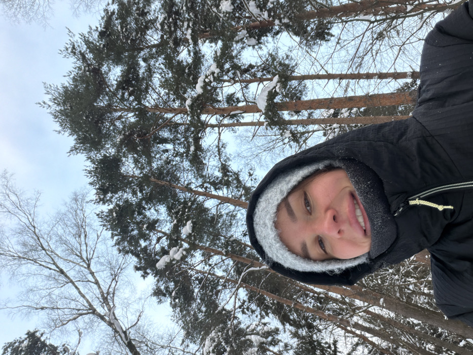 In Nemchinovo, Russia, a female individual is captured in an upward-angled selfie within a snowy wooded environment. The subject occupies the lower foreground, looking directly at the camera with a slight smile that reveals her teeth. She is attired in a light grey knitted beanie under a black, zippered hooded winter jacket, with visible snow or frost particles on the inner collar fabric around her face. The background is composed of a dense forest under a light blue-grey sky, featuring numerous tall pine trees with dark green, snow-dusted needles. Bare branches of deciduous trees, also lightly covered in snow, are visible, particularly in the upper left quadrant of the frame. The scene indicates a winter setting.