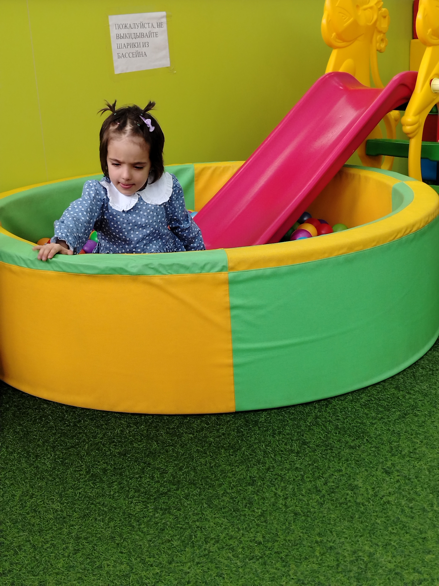 The image shows a young girl sitting in a ball pit with a slide. The ball pit has a green and yellow exterior and a pink slide. The girl is wearing a blue dress with a white collar. There is a sign on the wall in the background. The floor appears to be covered in artificial grass.