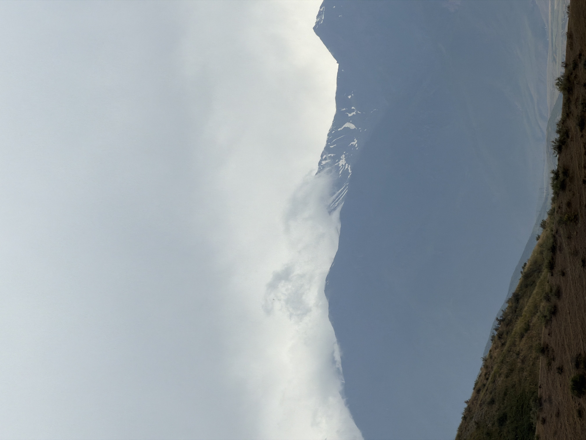 An overcast sky dominates the upper portion of the image, with a thin layer of clouds obscuring the sun. Beneath the clouds, a range of majestic mountains stretches across the frame. The peaks are shrouded in a soft, hazy light, with patches of snow visible on some of the higher elevations. The lower slopes of the mountains are covered in a dark, muted green foliage, suggesting a remote, natural landscape. In the foreground, a dry, brown hillside slopes gently downwards, dotted with sparse, scrubby vegetation and patches of bare earth. The overall atmosphere is one of serene, untouched wilderness, evoking a sense of calm and vastness. The scene is devoid of human presence and any signs of civilization, reinforcing the feeling of isolation and natural beauty.