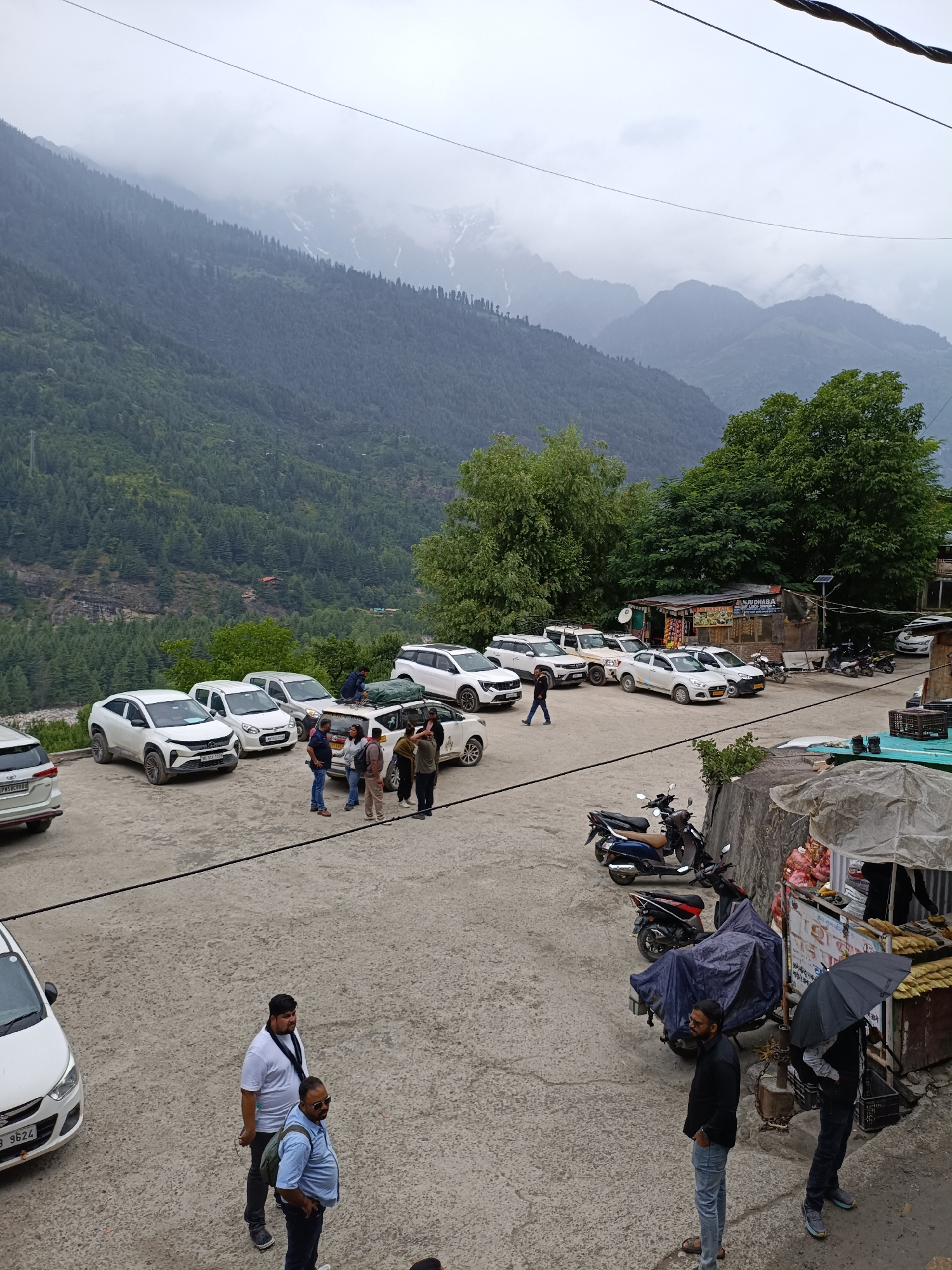 The image depicts a parking area and a roadside stall in Vashisht, India, with a mountainous, forested landscape in the background. In the foreground, several white SUVs and cars are parked. A group of men are standing and interacting near some of the vehicles. Further back, a roadside stall with a sign that reads "MANU DHABA" is visible, with more vehicles and motorbikes parked nearby. Two men are standing near the stall, one holding a black umbrella. The environment is overcast, with a road leading away from the parking area and a steep, tree-covered mountain rising in the distance. The overall scene suggests a stopover point or a small commercial area in a mountainous region.