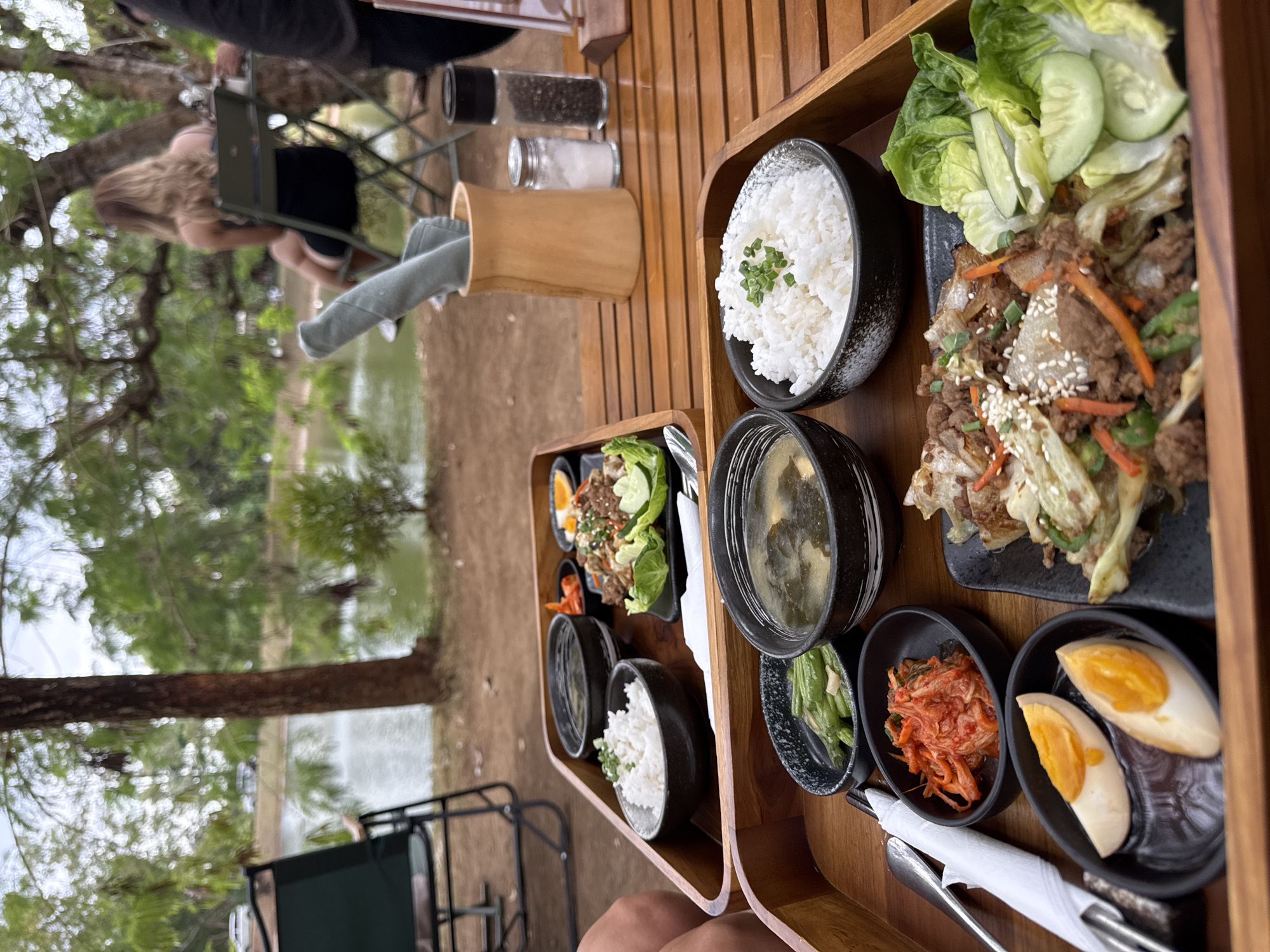 In Kuta Selatan, Indonesia, a wooden table outdoors hosts two wooden trays of Korean food, set against a backdrop of lush greenery and a calm body of water. The foreground tray features a bowl of rice topped with chives, a bowl of soup, green beans, kimchi, and sliced hard-boiled eggs. The second tray, slightly behind and to the left, holds rice, a small dish with meat and vegetables, and leafy greens with cucumber slices. A woman with blonde hair, wearing a black dress, sits at a table in the midground, facing away from the camera, with a wine glass in her hand. A folded gray napkin rests in a wooden holder on the table, beside salt and pepper shakers. A person's arm, wearing a dark sleeve, is visible on the far right. In the background, a tree with sparse leaves stands beside the water, with cars visible in the distance and another person seated at a table. A foldable chair is present in the far left.