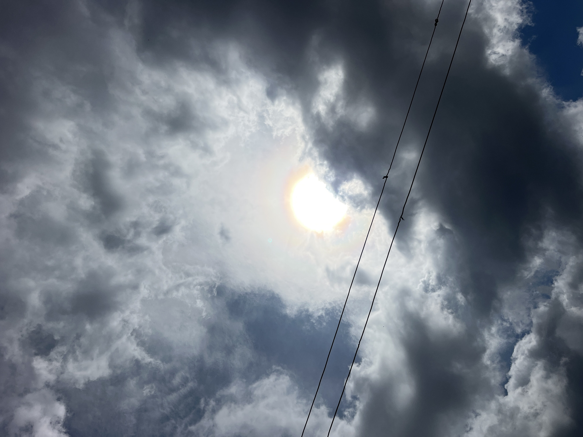 This is a low-angle shot of the sky, filled with dramatic, dark clouds. The sun is partially obscured by these clouds, creating a bright, almost glowing halo effect around it. The sun appears to be high in the sky, suggesting it's likely daytime, possibly late morning or early afternoon. Two dark, thin power lines stretch diagonally across the frame from the upper right to the lower left, interrupting the view of the clouds. The clouds themselves are varied in texture and darkness, with some appearing dense and stormy, while others are wispy and lighter, allowing glimpses of a bright blue sky in some areas. The overall mood is somewhat ominous due to the dark clouds, but the bright sun also offers a sense of light breaking through. There are no people or identifiable objects in the scene other than the power lines.