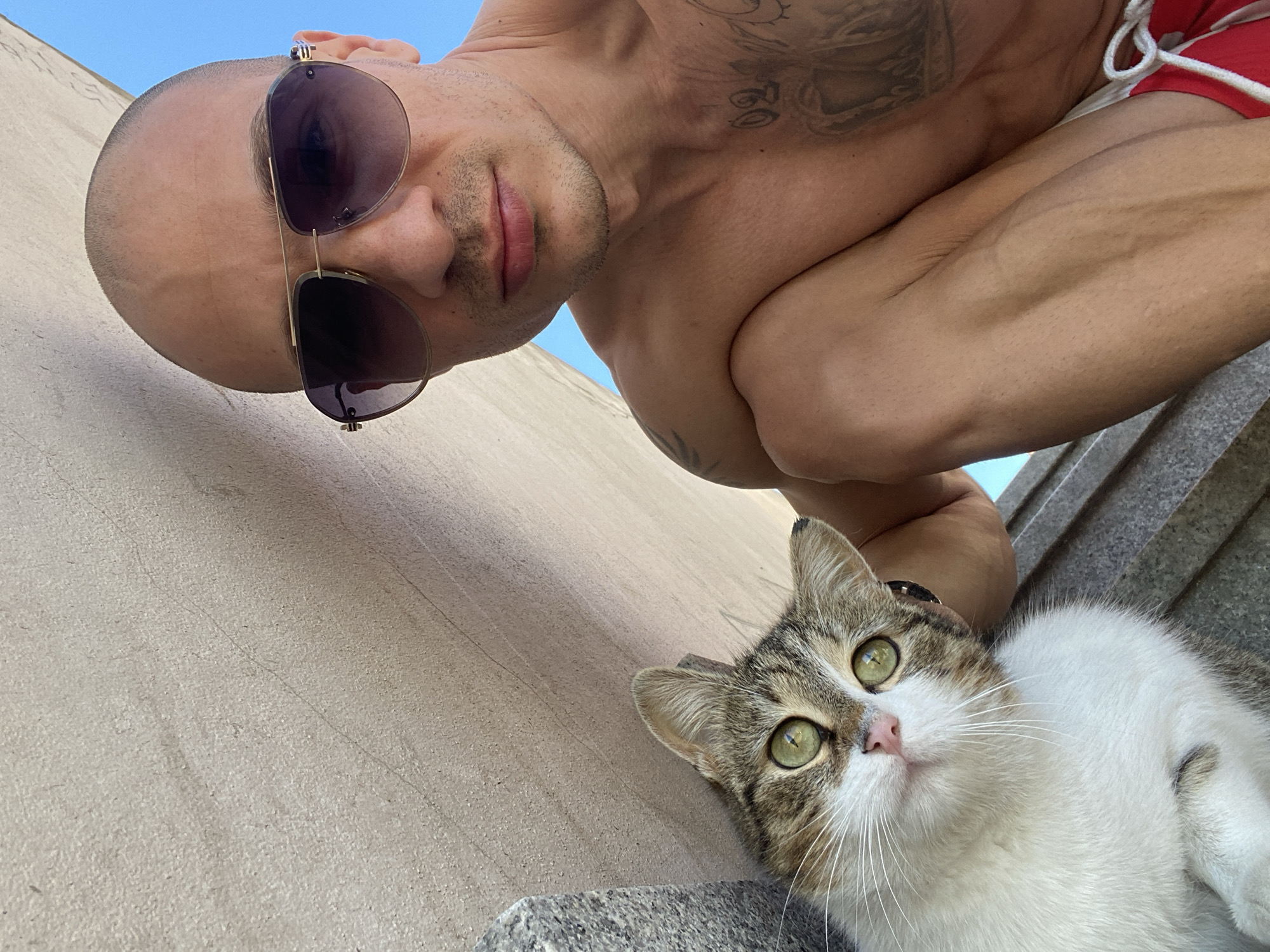 A man with a shaved head and sunglasses looks downward into the camera. He is bare-chested with tattoos visible on his upper chest and arms. A cat with green eyes and white and brown fur is situated in the bottom right corner of the frame, looking upwards. The background suggests a concrete or stone wall, with blue sky visible in the upper part of the frame. The scene is outdoors, likely in daylight. The city is Pleven, Bulgaria. The man and the cat are the primary subjects.
