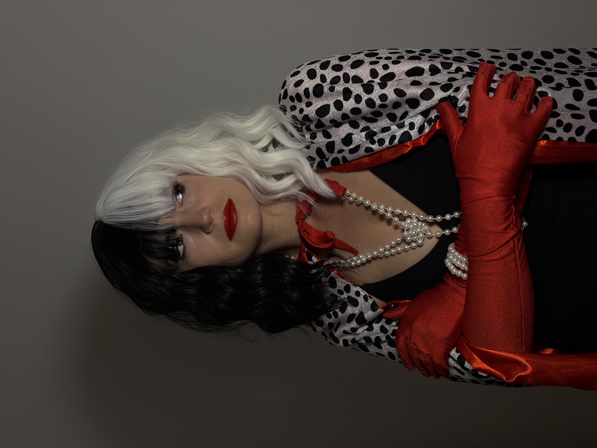 A person dressed as Cruella de Vil is positioned on her side with a gray background. She has a half-white and half-black wig, red lipstick, and is wearing a pearl necklace. She is wearing a black top and a Dalmatian-print shawl with red lining. Her hands are covered in long red gloves. The location is unknown and unidentifiable as the city of Budva, Montenegro.