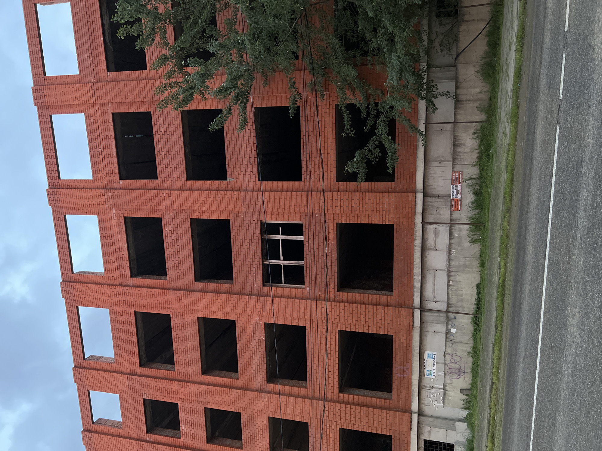 An eye-level, outdoor shot shows a portion of the exterior of an unfinished red brick building in the city of Yablonovskiy, Russia. The structure is about six stories high and features many rectangular windows without glass. The building is adjacent to a concrete retaining wall, a patch of grass, and a black asphalt road with a dividing white line. A leafy tree is partially visible in front of the upper-right part of the building. The sky is overcast.
