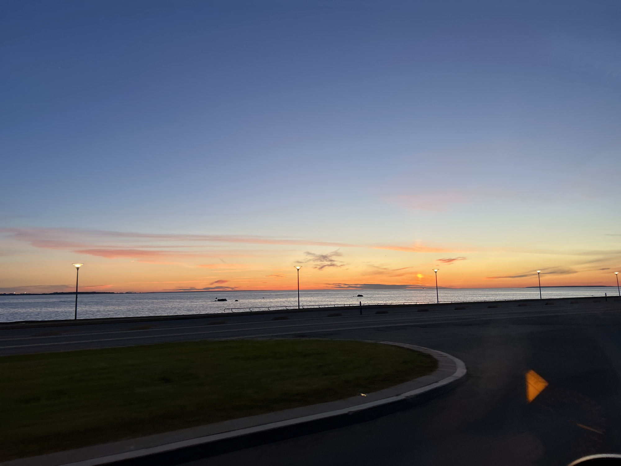 The image depicts a coastal scene in Tallinn, Estonia, during twilight. The sky transitions from a deep blue at the top to vibrant orange and pink hues near the horizon, where the sun is setting over the calm sea. Several illuminated lampposts are spaced along a paved walkway that runs parallel to the water. The sea is calm, with visible rocks and a distant landmass. In the foreground, a curved grassy area borders a dark asphalt road. The overall atmosphere is serene, with the natural light of dusk creating a peaceful ambiance.