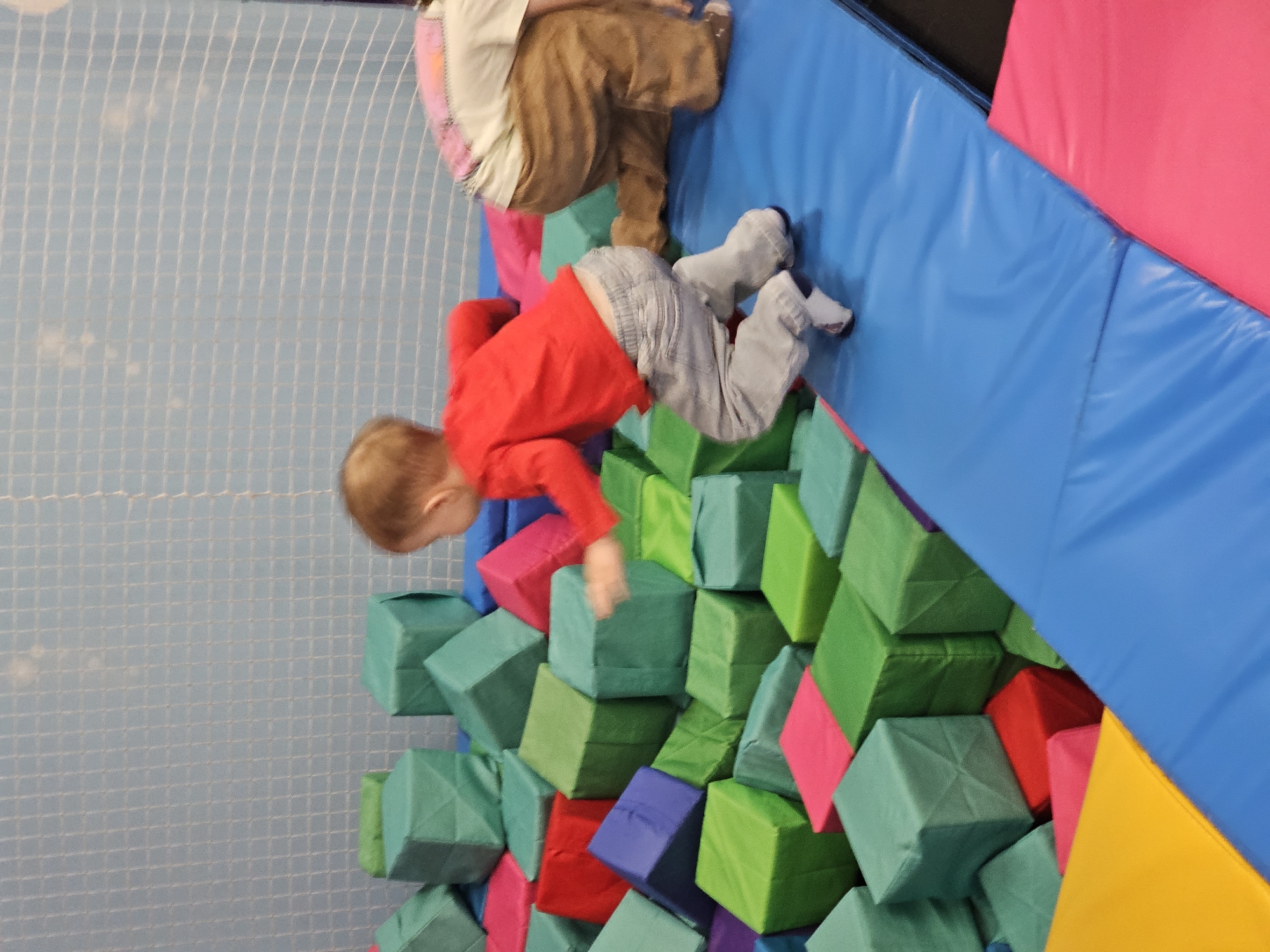 A child in a red shirt and grey pants is falling headfirst into a pile of green, blue, pink, and red foam blocks. Another child wearing brown pants is climbing on a blue padded wall. The background includes a blue grid wall, and a padded wall with pink and yellow sections. This scene appears to be in a play area or gym in Khimki, Russia.