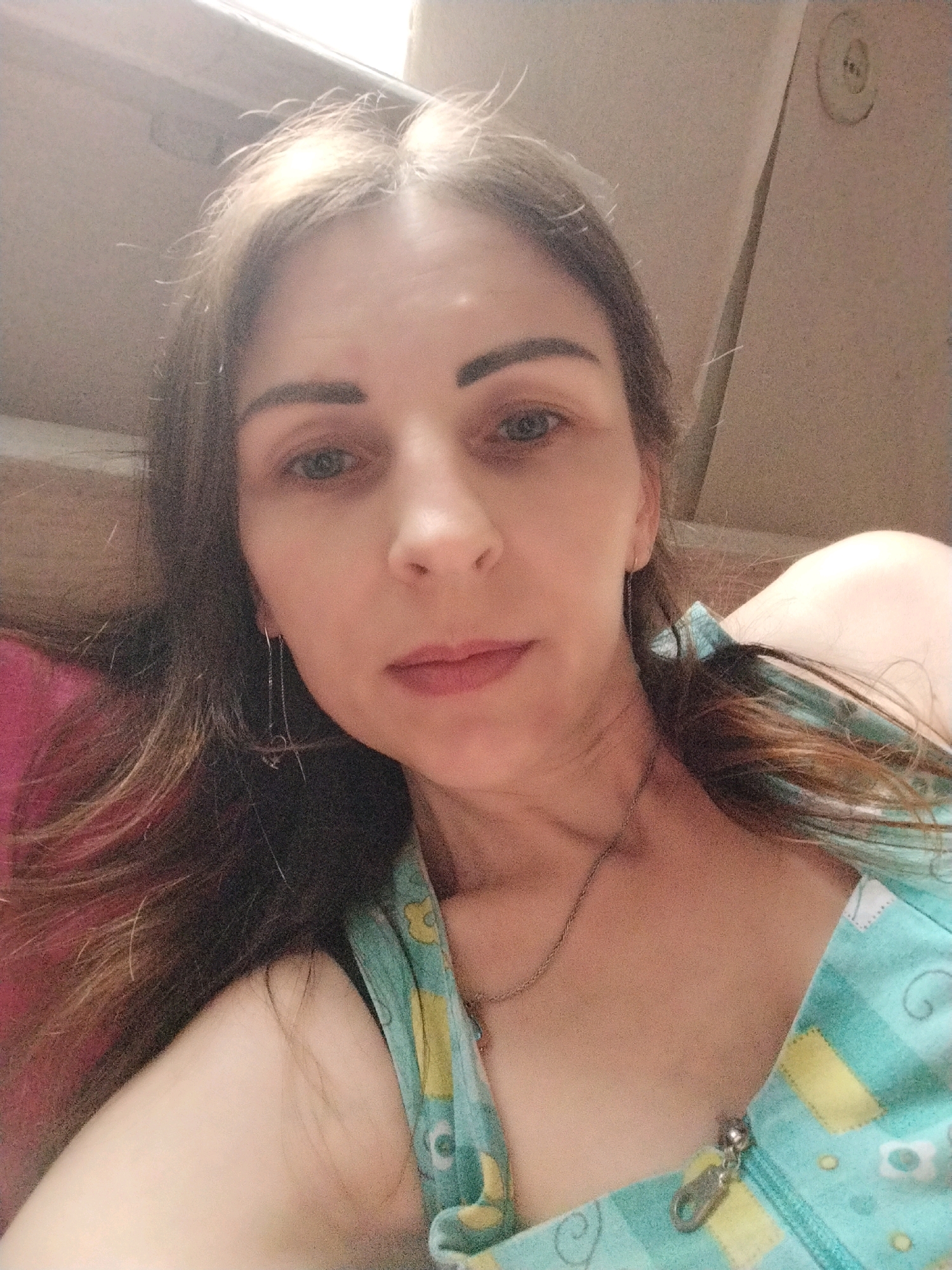 This is a close-up selfie of a woman with long, brown hair and blue eyes. She is wearing a light blue and yellow patterned top with a zipper closure. She has on a silver chain necklace and long, dangling earrings with small charms. The background appears to be an indoor setting with a beige wall and a window or light source at the top. There is no visible indication of the city of Vinnytsia, Ukraine in the image.