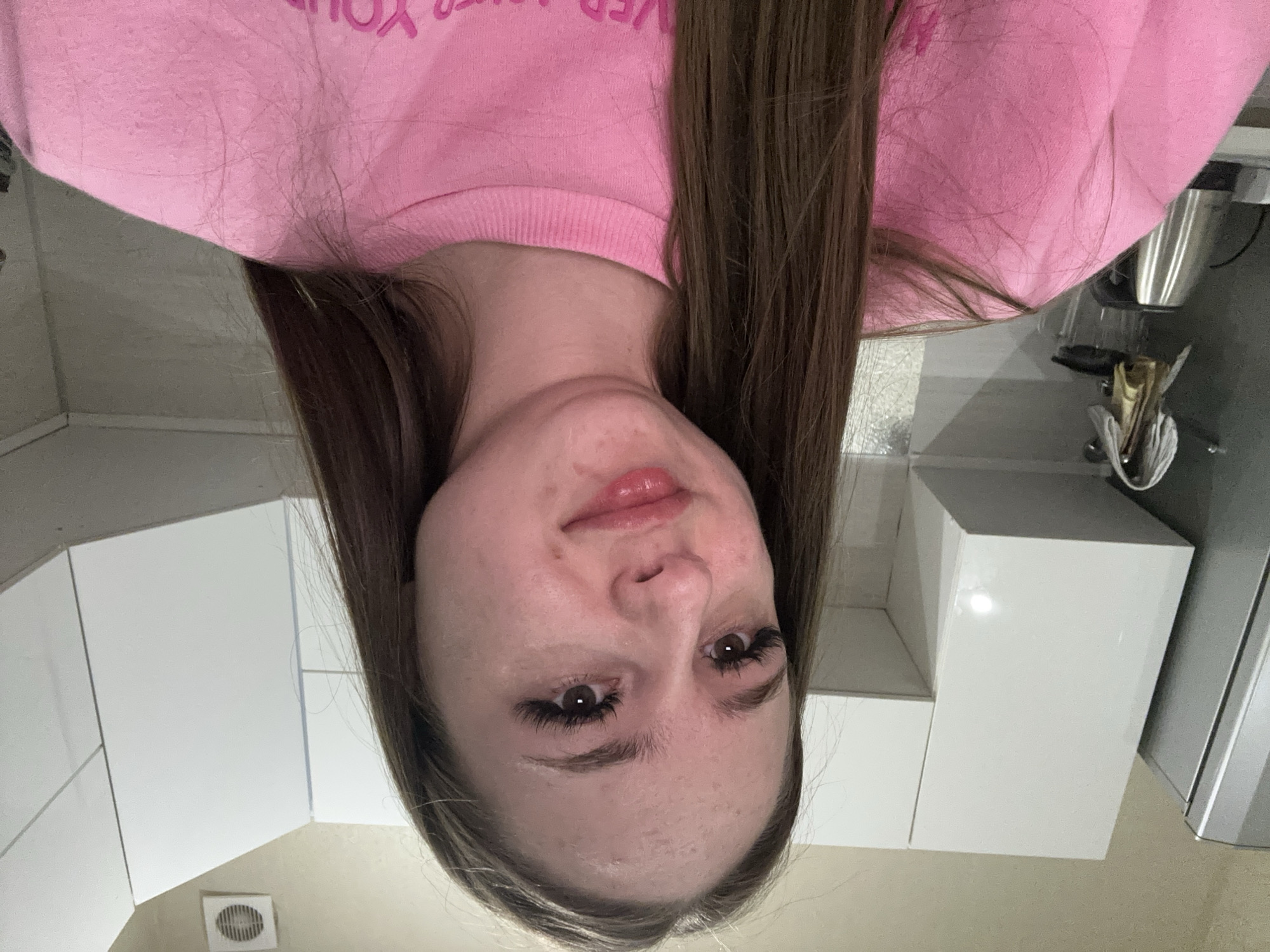 The media file shows a portrait of a young woman with long, brown hair. She is wearing a pink sweatshirt with text on it. Her face is tilted upwards, and she has makeup on, including mascara and lip gloss. Her eyes are open, and she is looking directly at the camera. The background appears to be a kitchen or bathroom setting, with white cabinets visible. There is also a stainless steel appliance and some shelving in the background. The image is taken from a low angle, making the woman appear to be looking down. The lighting suggests it is daytime, but the specific time of day is not clear. There is no discernible weather information. The overall emotion conveyed by the woman is neutral or possibly slightly curious. The text on her sweatshirt is partially visible and appears to be in a bright pink color.
