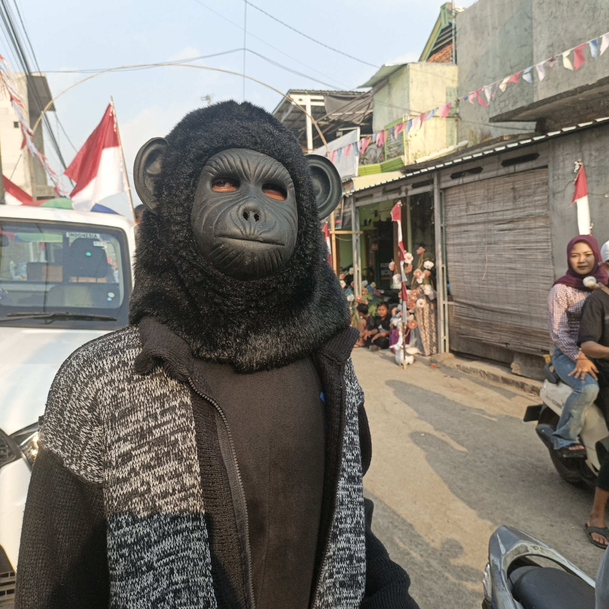 In Tanahbaya, Indonesia, a person is depicted wearing a black monkey mask and a grey and black knitted jacket over a black shirt. The individual is standing in front of a white vehicle. In the background, there are buildings decorated with small Indonesian flags and pennant banners. Several people are visible in the distance, including a woman wearing a headscarf and a plaid shirt sitting on the back of a motorcycle, and other individuals seated on the ground. The scene appears to be outdoors during the day, with shadows cast on the street, suggesting a street-level view of a community gathering or event.