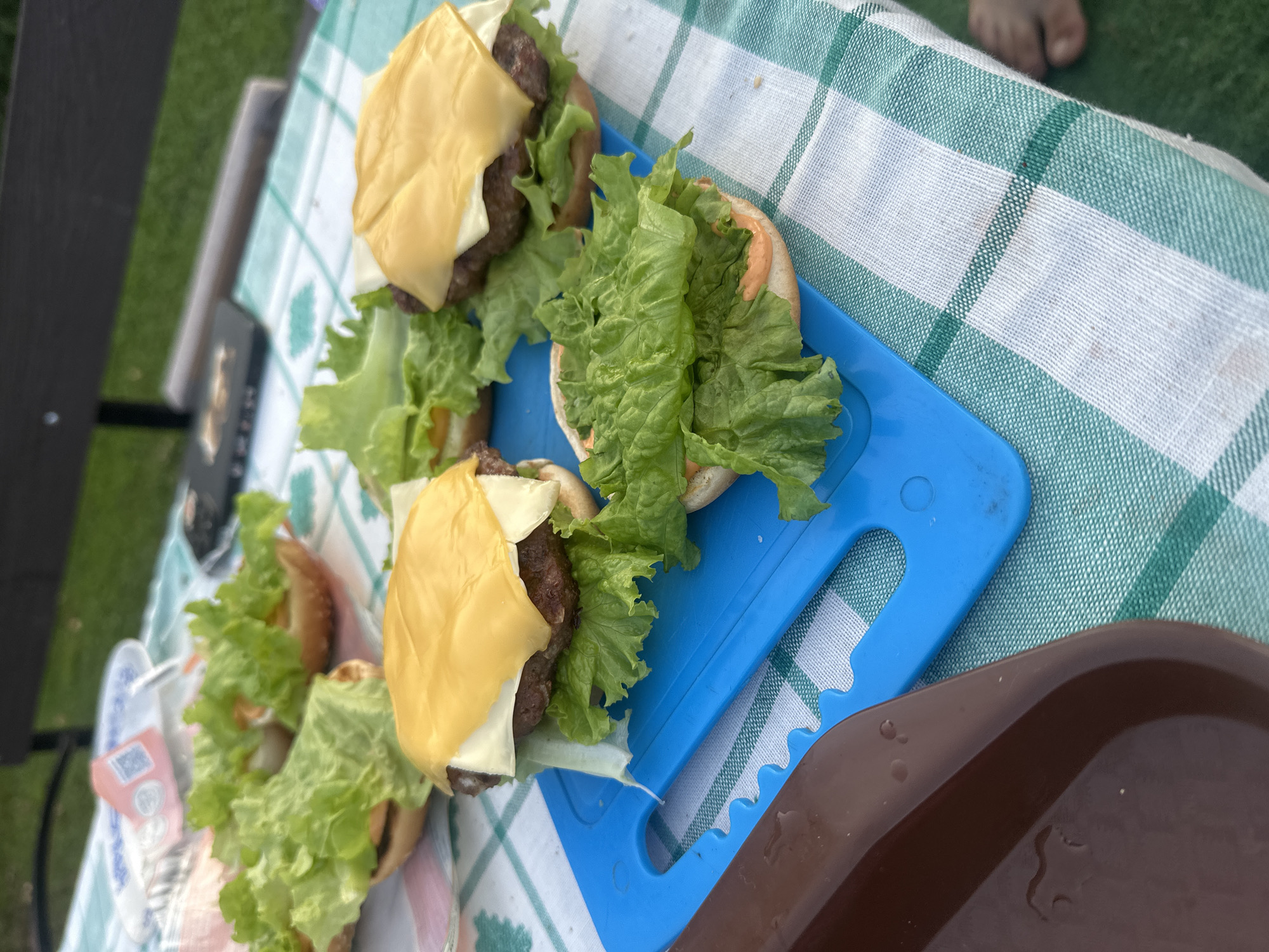 This is a close-up, high-angle, outdoor shot of several cheeseburgers being assembled on a picnic table covered with a green and white checkered tablecloth. The scene is set in Galewo, Russia. In the foreground, a blue plastic cutting board holds two partially assembled burgers. One burger on the board has a beef patty topped with lettuce and a slice of melted yellow cheese. The other burger on the board is the bottom half of a bun with a generous serving of lettuce and what appears to be a pinkish sauce. Behind the cutting board, other assembled burger halves are visible, some with lettuce and others with a patty and cheese. In the background, a dark-colored park bench is partially visible, and the scene appears to be set on a grassy area. The lighting suggests it is daytime.