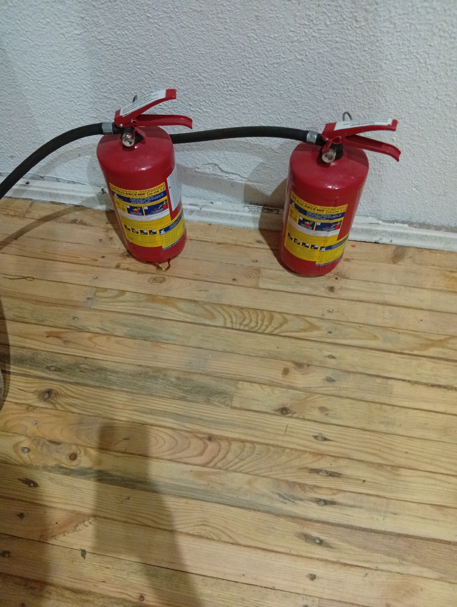 Two red fire extinguishers with black hoses are placed on a wooden floor against a textured white wall. The fire extinguishers have red handles and labels with Cyrillic text and diagrams. The floor is made of light-colored wooden planks with visible grain and knots. A shadow of a person is cast on the floor to the right of the fire extinguishers. The scene appears to be indoors, likely in a storage area or room. There is no visible indication of the specific location within Крыўлянскі сельскі Савет, Belarus.