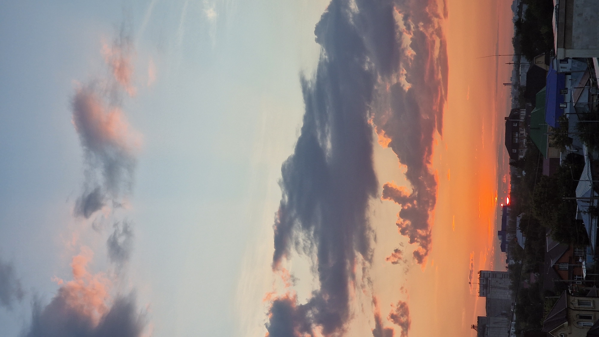 The image depicts a sunset over a city skyline, likely in Kostanay, Kazakhstan. The sky is a gradient of colors, transitioning from pale blue at the top to warm orange and pink hues near the horizon. Fluffy, dark gray clouds are scattered across the sky, some tinged with pink from the setting sun.
Below the sky, a low-rise urban landscape is visible. The foreground features rooftops of various houses, some with visible chimneys. A street light is present in the lower portion of the image. Further in the background, there are trees, smaller buildings, and a few larger industrial structures, including what appears to be a grain elevator or silo complex on the left. The sun itself is a bright orange orb, partially obscured by clouds and buildings on the horizon. The overall atmosphere is calm and serene, characteristic of a late evening. No people are visible in the frame.