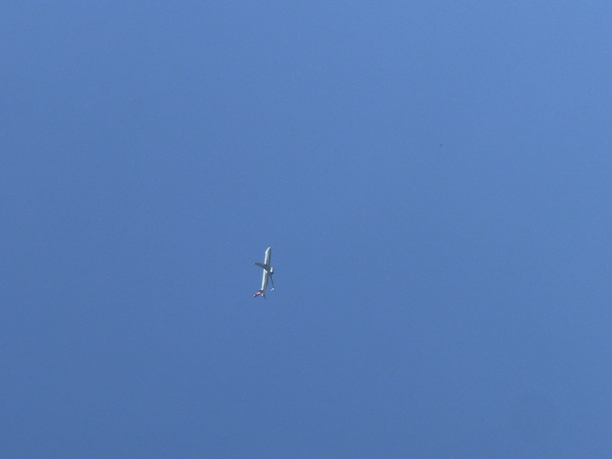 A white commercial airplane with a distinct red marking on its tail is captured flying against a clear, bright blue sky. The aircraft, appearing to be a twin-engine passenger jet, is seen from below, positioned in the upper-middle section of the frame. Its orientation is diagonal, suggesting it is either ascending, descending, or banking in flight. The sky is completely free of clouds, indicating fair weather and bright daylight conditions, which illuminate the underside of the plane. No other objects, landscapes, or discernible text are visible in the image. The scene depicts an airplane in transit over what is contextualized as Varna, Bulgaria.
