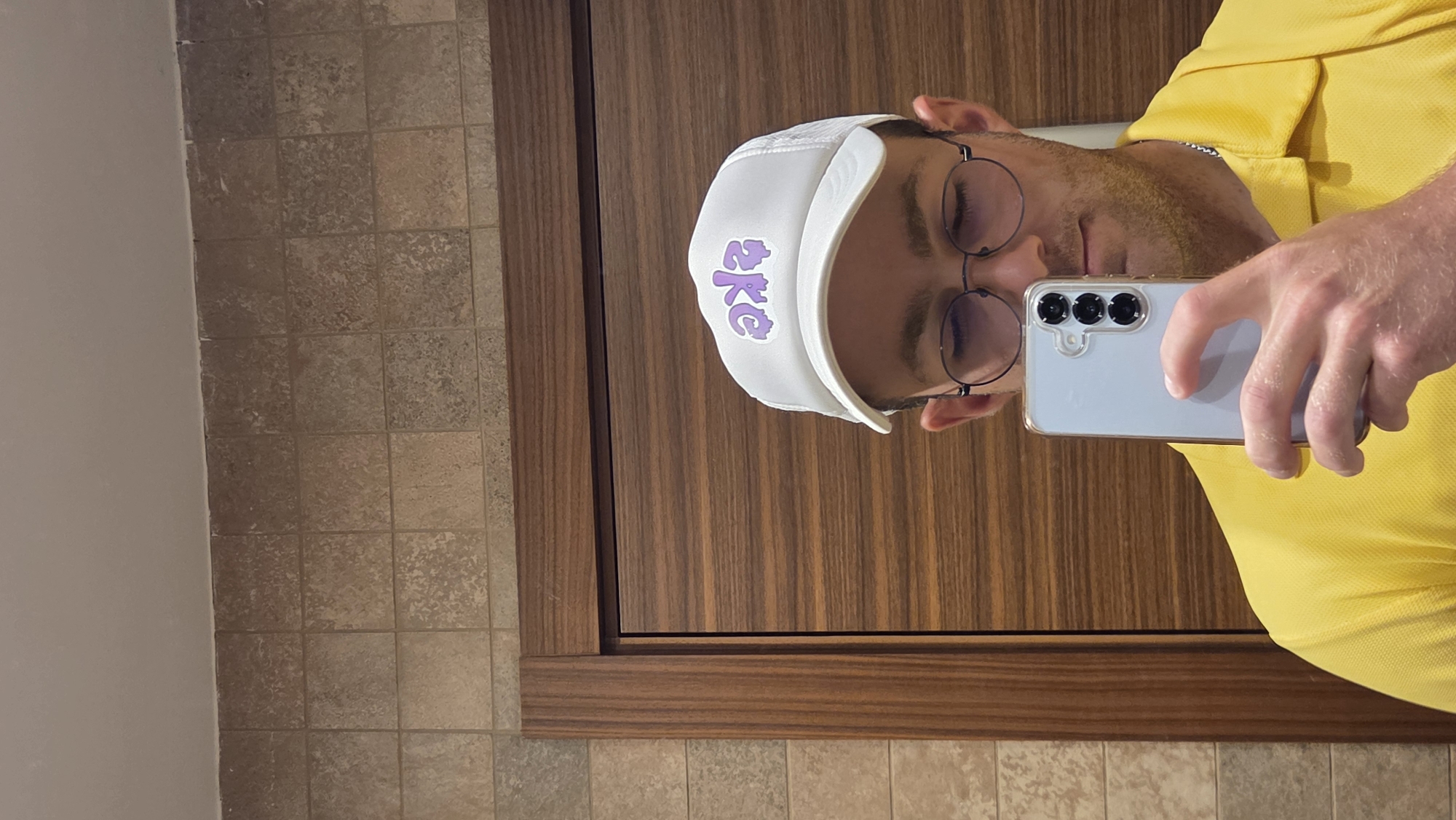 A man, wearing glasses, a white trucker hat with purple lettering, and a yellow collared shirt, takes a selfie in a bathroom. The background features tiled walls and a wooden door. The man is holding a light blue smartphone in his right hand, angled to capture his reflection.