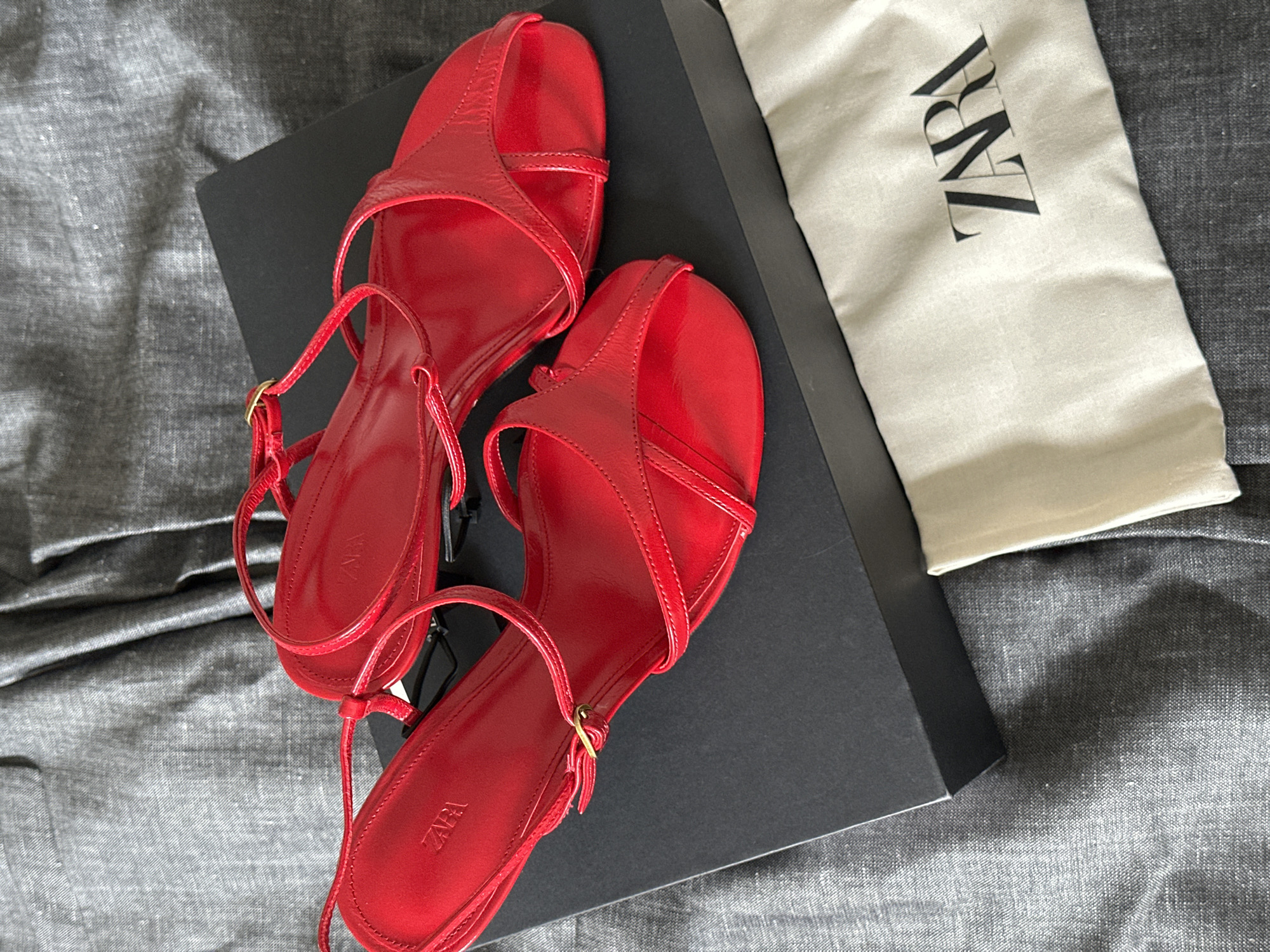 This media file contains an overhead shot of a pair of bright red, open-toe sandals. The sandals are made of a shiny material and feature thin straps that crisscross over the foot and connect to a thin ankle strap with a gold buckle. The shoes are placed on top of a black shoebox. To the right of the shoes and box, there is a light-colored fabric dust bag with the black "ZARA" logo printed on it. The items are situated on a textured gray surface, possibly a bedspread or fabric backdrop, which appears to be slightly rumpled, suggesting a casual indoor setting. There is no activity or event taking place; the image is a static product shot. The lighting suggests it might be daytime, but no specific time of day can be determined. There are no people, weather, or emotional cues visible. The visible text is "ZARA" on the dust bag and discreetly on the insoles of the sandals.