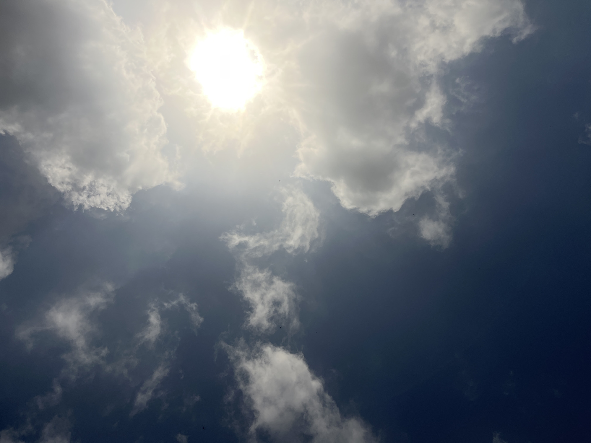The image shows a view of the sky, likely taken during the day. The sun is visible, partly obscured by clouds. The sky's color ranges from bright near the sun to a darker blue away from it. Some clouds are white and puffy, while others appear more diffused. The angle of the shot suggests the camera was pointed upwards. It is impossible to confirm the location is Ikot Ekpene, Nigeria, with only visual data. There are no people or objects visible in the image other than the sky and clouds.