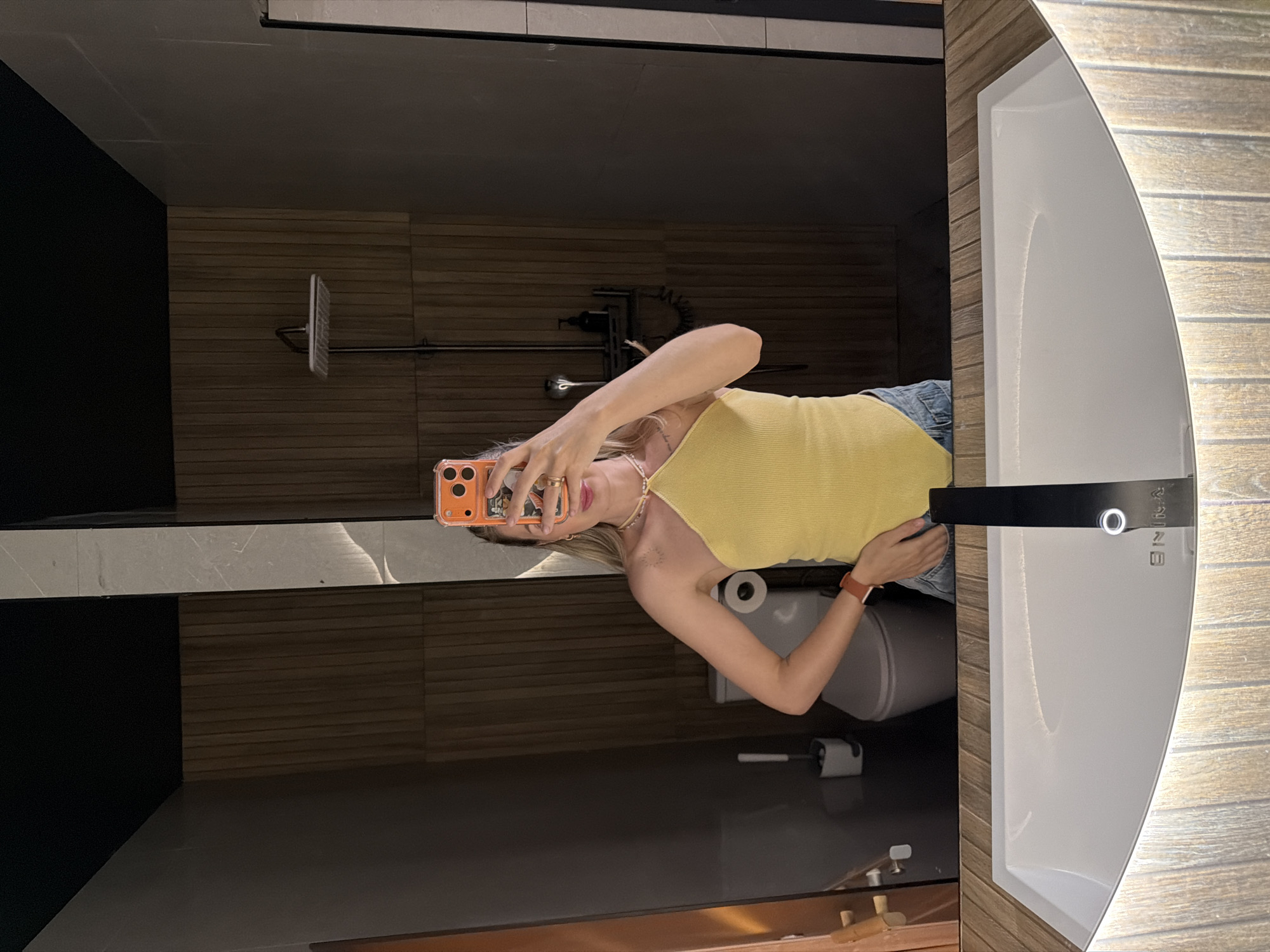 A woman is taking a selfie in a bathroom. She is wearing a yellow halter top and blue jeans. The bathroom has wood-paneled walls, a toilet, and a shower. The sink is in the foreground, with a black faucet. The image is taken from the chest up. The woman is holding an orange smartphone in her right hand, which is angled to capture her reflection in the mirror. She has blonde hair and is wearing jewelry. The background shows a toilet and a showerhead. The environment appears to be a modern, well-lit bathroom. The city of Семилуки, Russia is mentioned, but there are no visual cues in the image to confirm this location.
