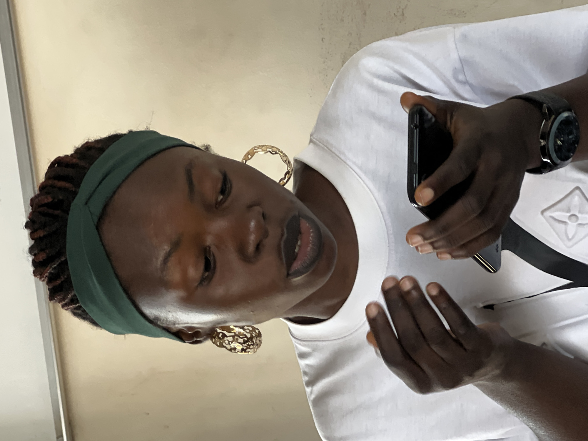 A dark-skinned young woman with dark braided hair styled with a green fabric headband and large gold hoop earrings is looking down at a smartphone she is holding in her hands. She is wearing a white t-shirt and a watch on her left wrist. The background is a plain, light-colored wall. The image appears to have been taken indoors, possibly in Jos, Nigeria.