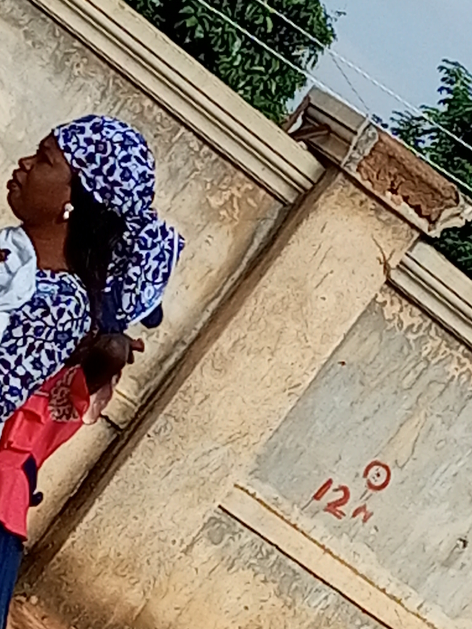 In Gombe, Nigeria, a woman wearing a blue and white patterned headscarf and earrings is seen in profile, looking upwards. She is draped in a white garment with a similar blue and white pattern, and a bright pink garment is partially visible around her neck and shoulders. The background consists of a textured, light-colored wall, possibly part of a building. Above the wall, green foliage from trees is visible against a pale blue sky. A number "12n" is written in red on the wall in the lower right quadrant of the image, with a small red circle next to the "n". The overall impression is a candid outdoor shot.