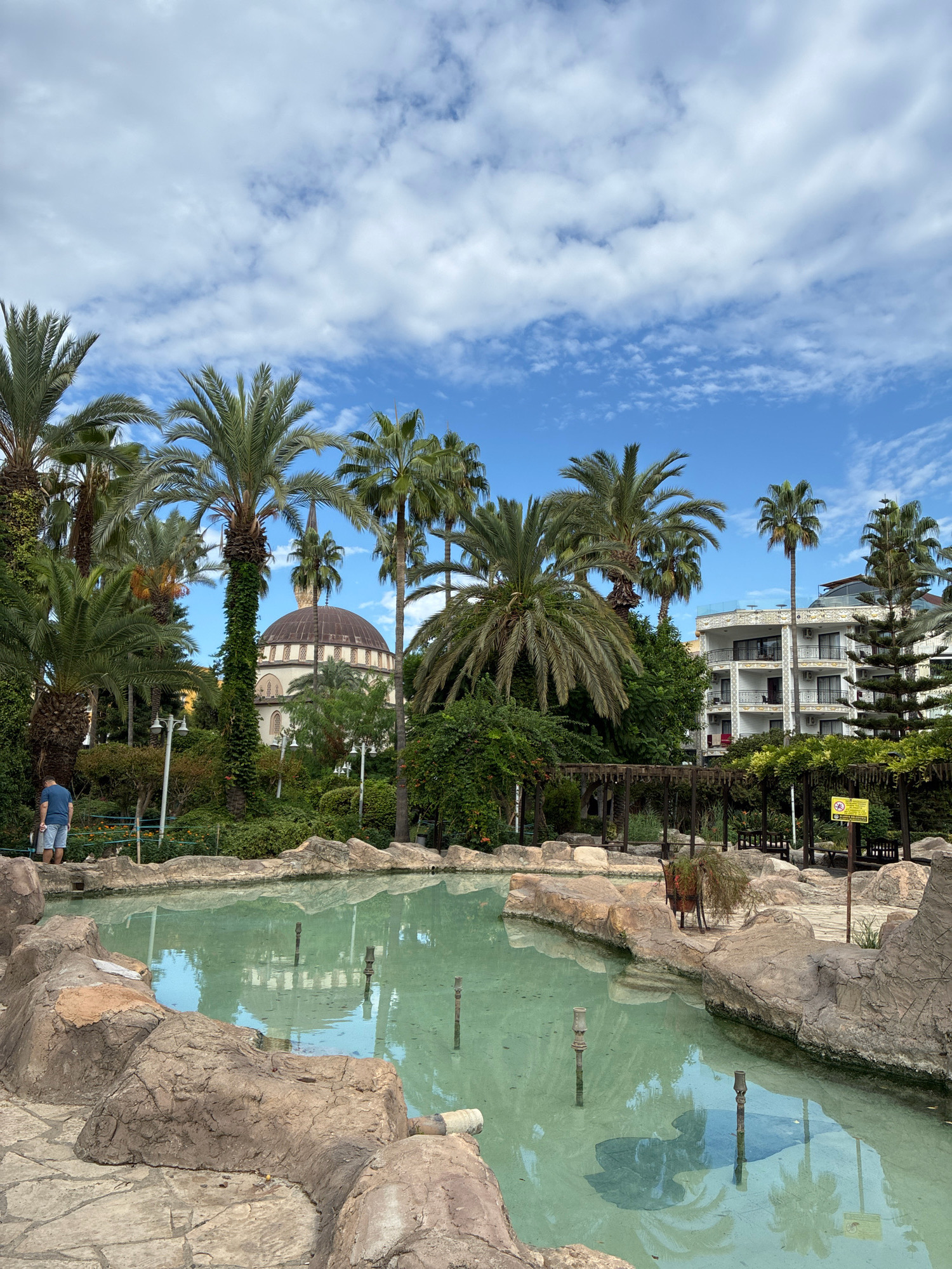 In Alanya, Turkey, a serene pond with clear turquoise water reflects the surrounding environment under a partly cloudy sky. The pond is framed by rugged, rocky edges and paved walkways. Palm trees, lush green bushes, and other foliage create a tropical ambiance.
In the background, a distinguished mosque with a large dome and a tall minaret stands amidst the greenery, suggesting a cultural or historical landmark. To the right, a modern hotel building with multiple balconies and white-tiled exteriors is visible, indicating a blend of old and new architecture.
A lone man in a blue t-shirt and shorts stands near the pond, facing away from the camera, observing the scenery. The pond itself appears to be a decorative water feature, possibly a dried-up fountain, with several spouts visible beneath the water's surface. Wooden pergolas with benches offer shaded seating areas, hinting at a public park or recreational space. A yellow warning sign is posted on a post near one of the pergolas. The overall scene evokes a sense of tranquility and a holiday atmosphere.