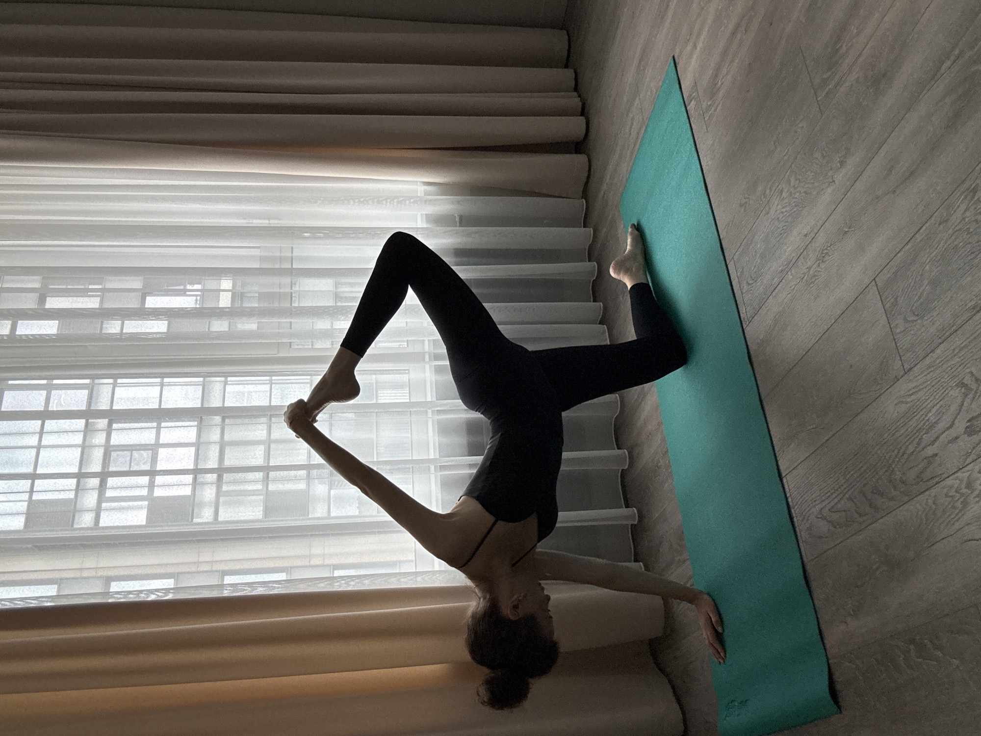 A person is practicing yoga on a green mat in front of a window with sheer and opaque curtains. The person is in a black leotard and leggings, with their hair in a bun. Their left knee is on the mat, and their right leg is extended upwards and bent, with the right hand holding the right foot. The left hand is on the mat, extended forward for support. The background shows a large window with multiple panes and an urban landscape visible through the sheer curtains. The floor is made of grey wood planks.