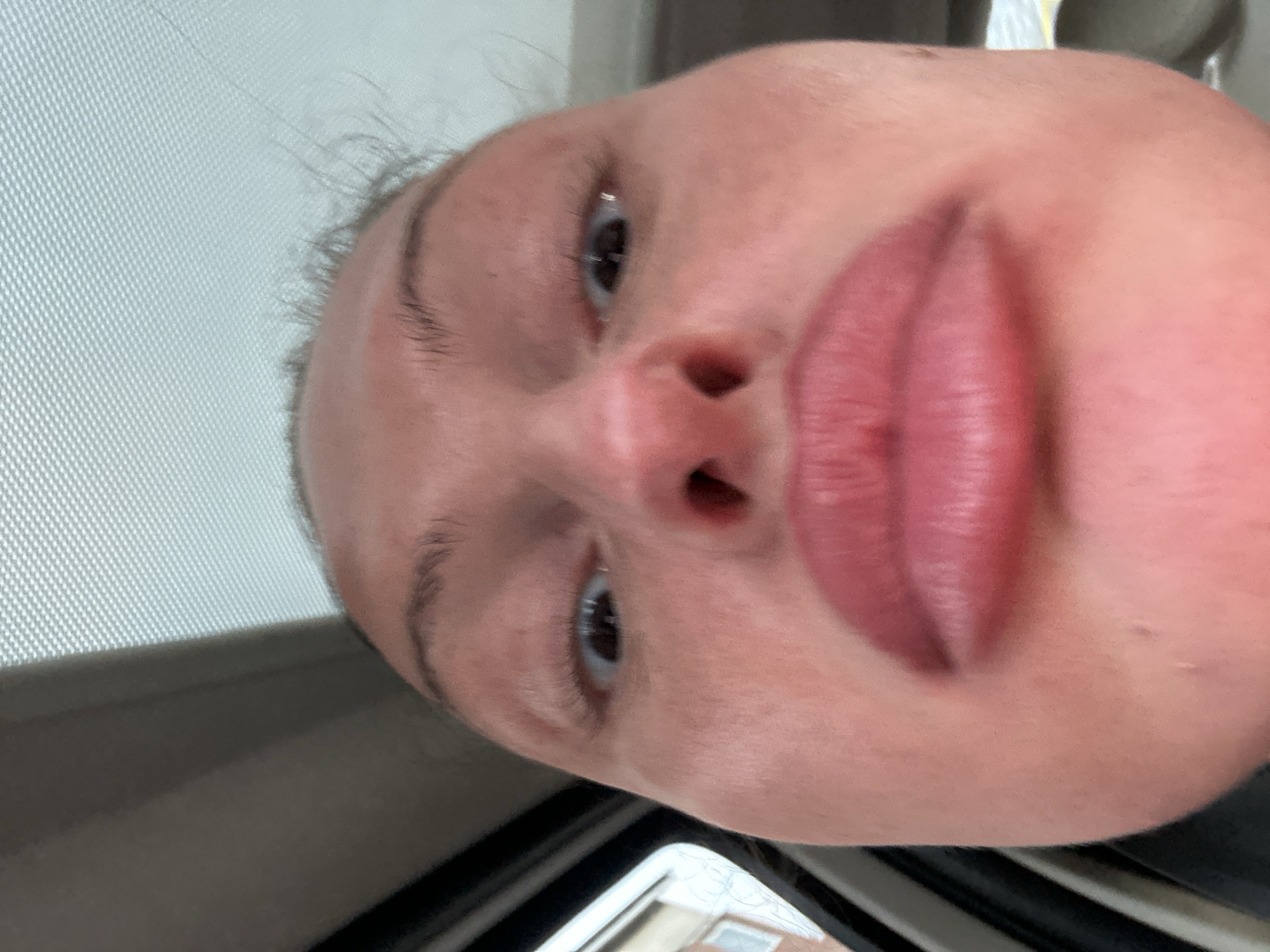 A close-up, low-angle view of a person's face, possibly female, is centered in the frame. The face is oriented at an angle, with the forehead at the top left and the chin at the bottom right. The person has fair skin, brown eyes, and full, somewhat reddened lips. The nose is prominent and centered. Eyebrows are visible but sparsely defined. The background shows a car sun visor with a mesh texture in the top left and a blurry, out-of-focus view of buildings through a car window at the bottom. The setting appears to be inside a vehicle. There is no evidence indicating that this picture was taken in Tonbridge and Malling, United Kingdom.