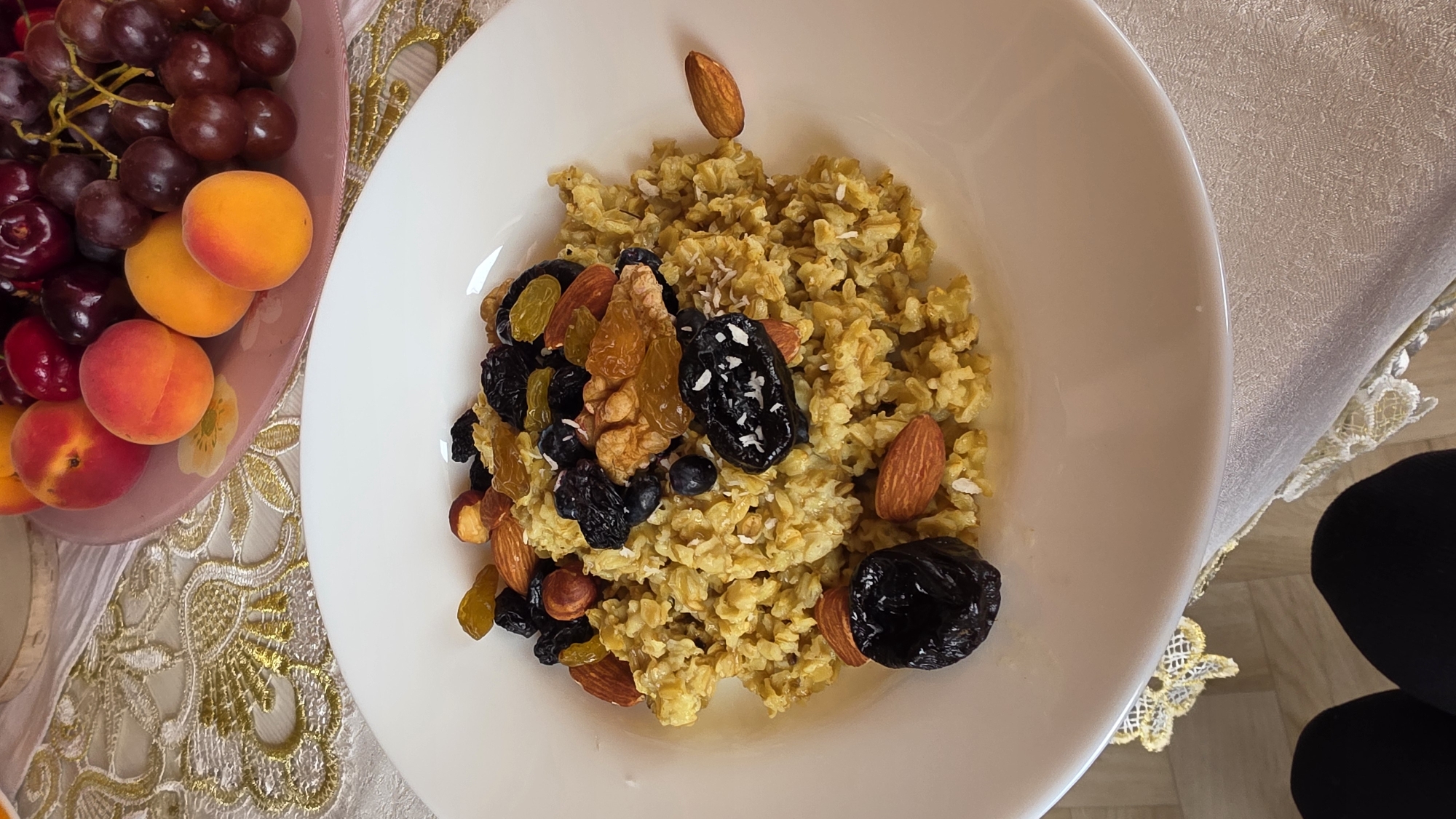 A high-angle, close-up shot captures a white bowl filled with oatmeal topped with various dried fruits and nuts. The oatmeal has a grainy texture and is a light brown color. Scattered across the top are plump, dark raisins, chopped almonds, a few whole hazelnuts, blueberries, and two large, dark prunes, one of which is sprinkled with shredded coconut. A single almond rests on the rim of the bowl. In the background, out of focus, is a pinkish-purple plate holding apricots and dark red grapes. The setting appears to be a table covered with a light-colored tablecloth featuring a gold embroidered pattern. The bottom of the frame shows the black leggings of a person seated at the table. The image does not contain any explicit information to determine the location within the city of Kupoyarovo, Russia.