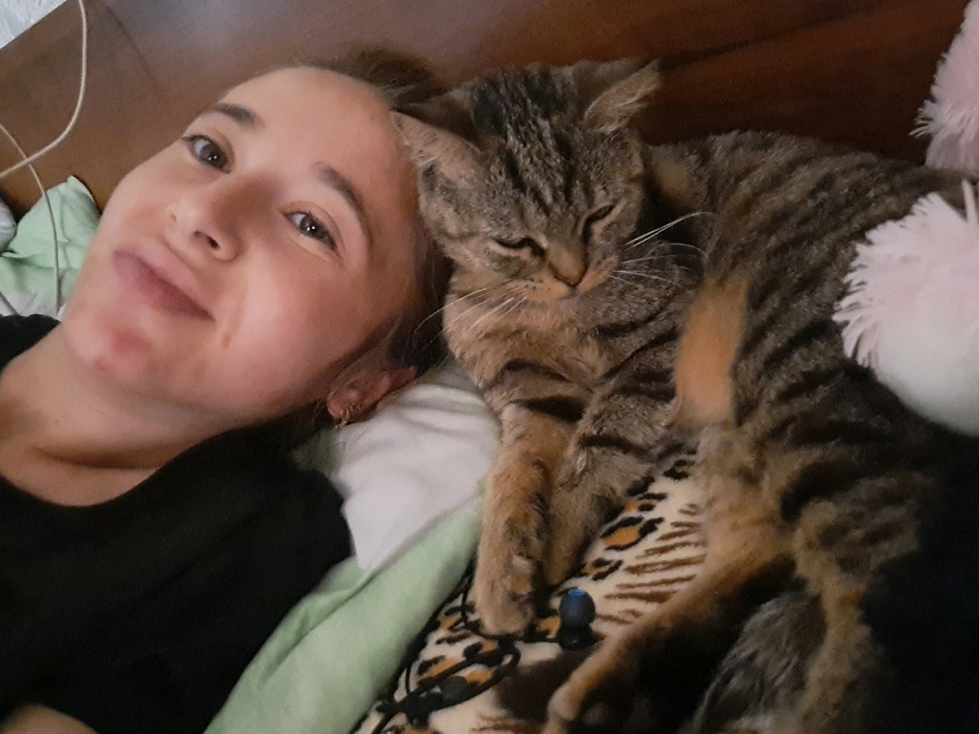A selfie taken in Vypasne, Ukraine shows a young woman with light skin and dark hair lying down and looking at the camera. She is wearing a black t-shirt. Next to her, a tabby cat is also lying down, with its eyes closed and appearing to be asleep. The cat is resting on a blanket with a leopard print pattern. The background is dark and indistinct, suggesting an indoor setting, possibly a bedroom. A white cord runs from the upper left corner of the image.