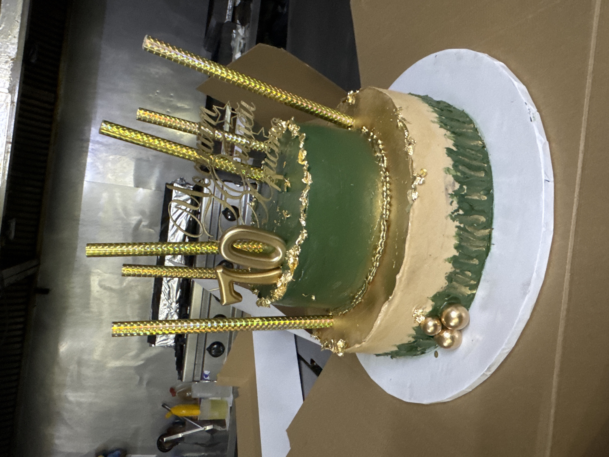 The image shows a two-tiered cake. The top tier is green and the bottom tier is a tan color. Both tiers are decorated with gold accents, including gold leaf and gold piping. There are gold candles on the cake, with a "70" topper. There is also a gold script cake topper that reads "Meghan Ruci". The cake is on a white cake board, which is placed on a cardboard surface. In the background, there is a stainless steel commercial kitchen with cooking equipment. The scene appears to be indoors in a kitchen or catering setting. The location is not specified in the image.